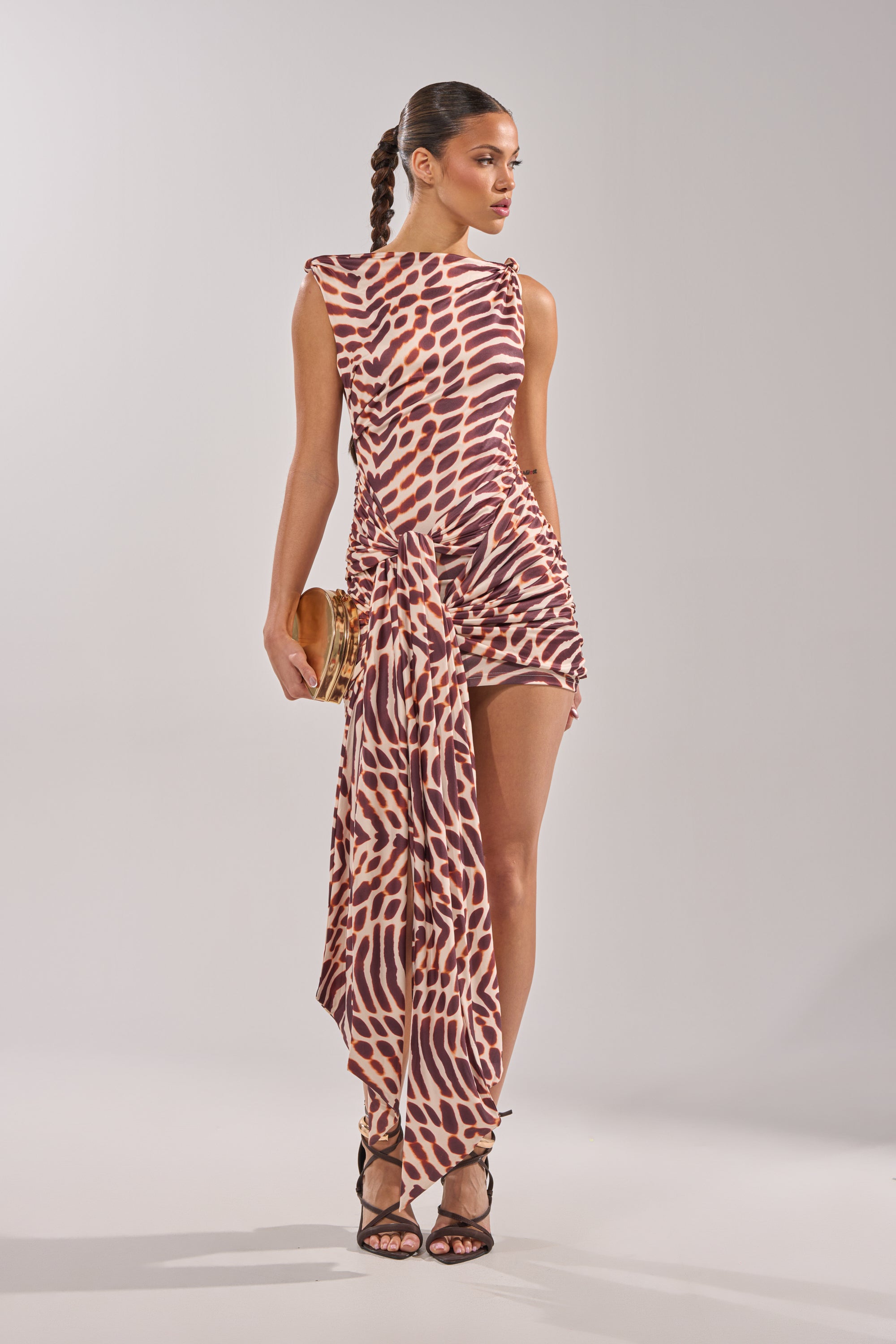 A woman in the JUNGLE TIES LEOPARD MINI DRESS—an off-the-shoulder, asymmetrical brown and beige animal-print mini dress—stands against a light gray background, holding a small clutch and wearing black strappy heels with her hair in a long braid. 2