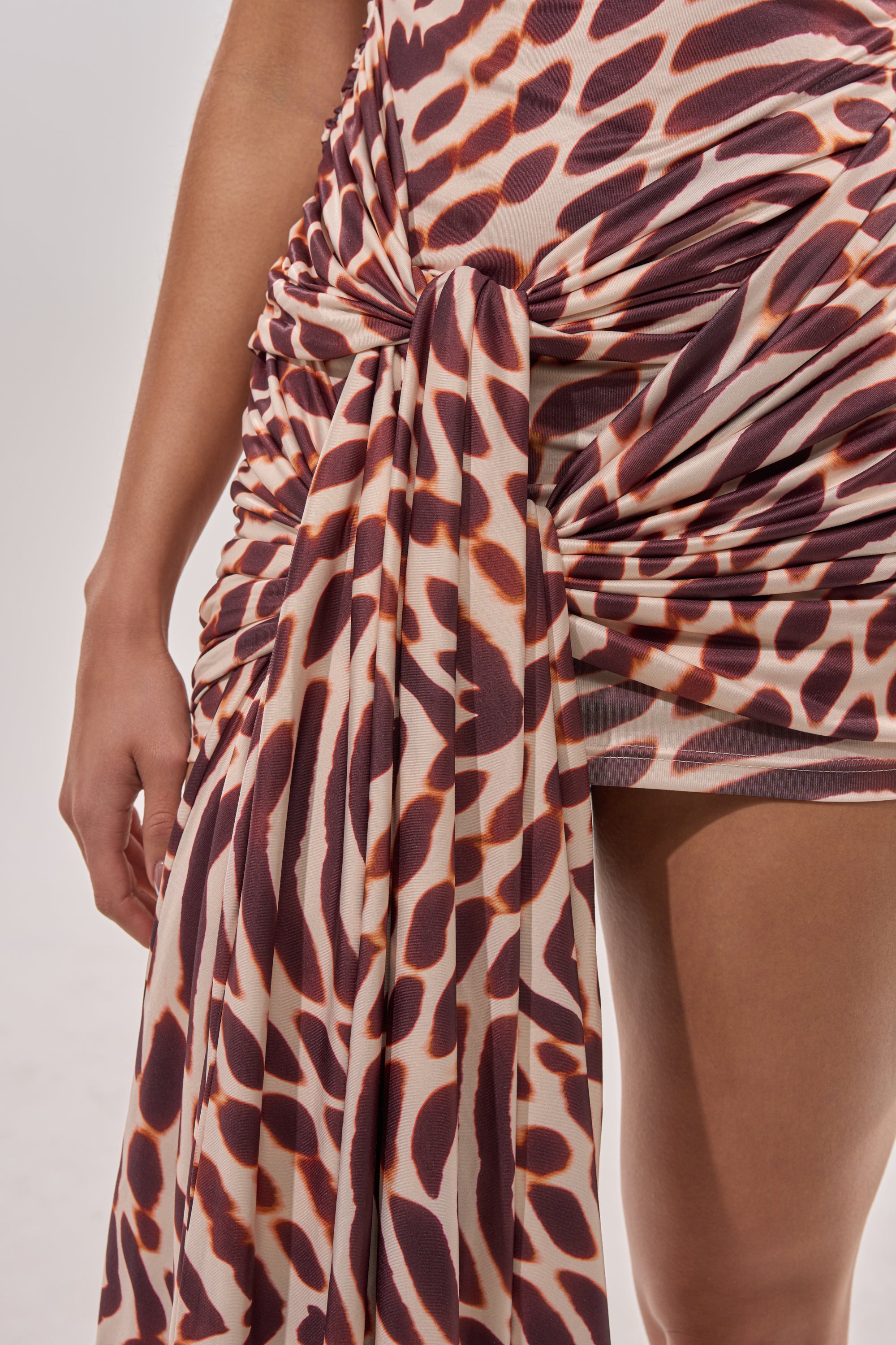 A close-up of a person in the JUNGLE TIES LEOPARD MINI DRESS, featuring bold brown and cream animal print, ruching, and a large tied detail at the hip. The person's arm and upper thigh are visible. 11