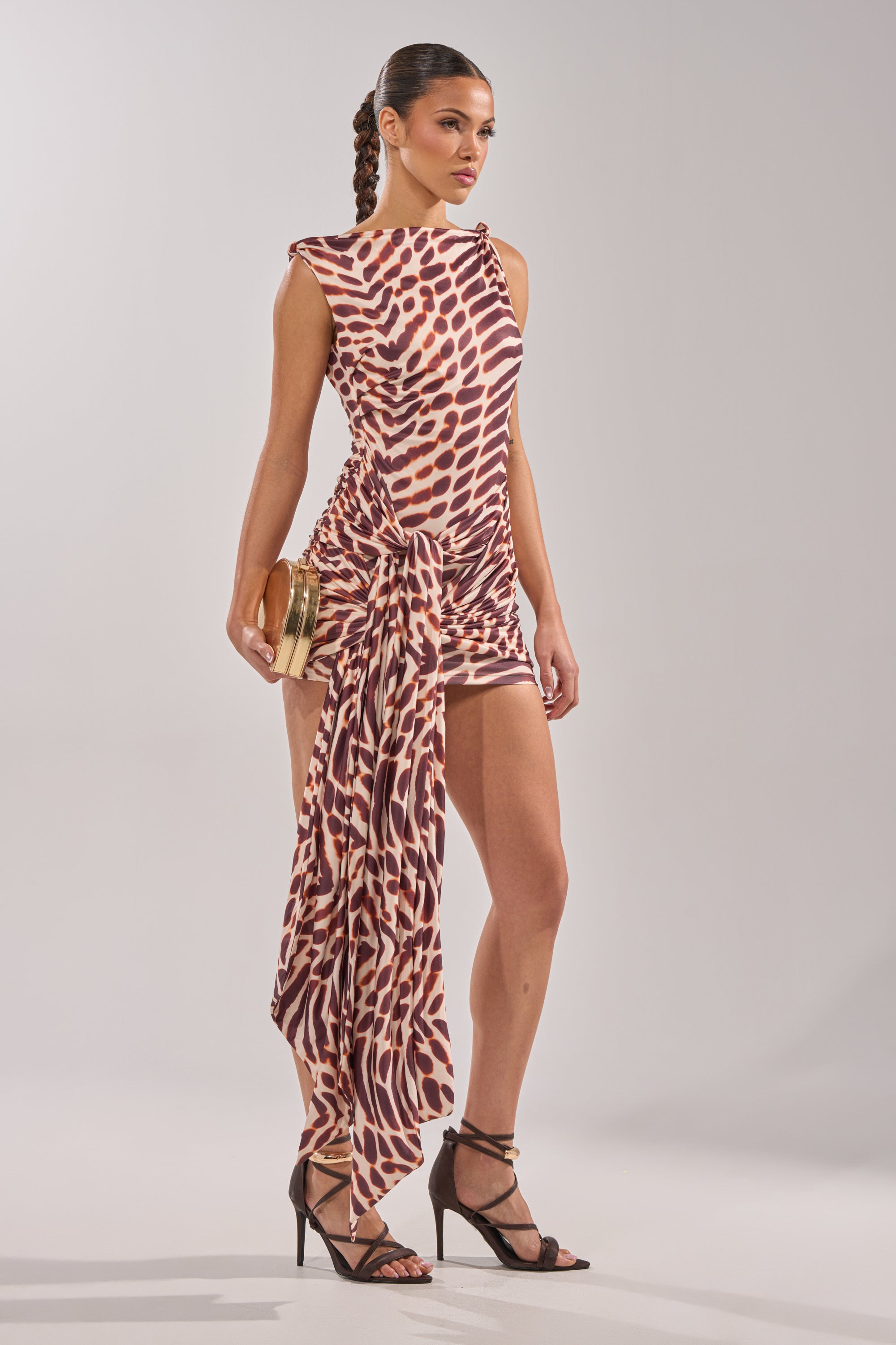A woman wears the JUNGLE TIES LEOPARD MINI DRESS, a sleeveless asymmetrical mini with brown and beige leopard print, ruched detail, and a long draped side. She holds a gold clutch and wears black heels against a plain background. 3
