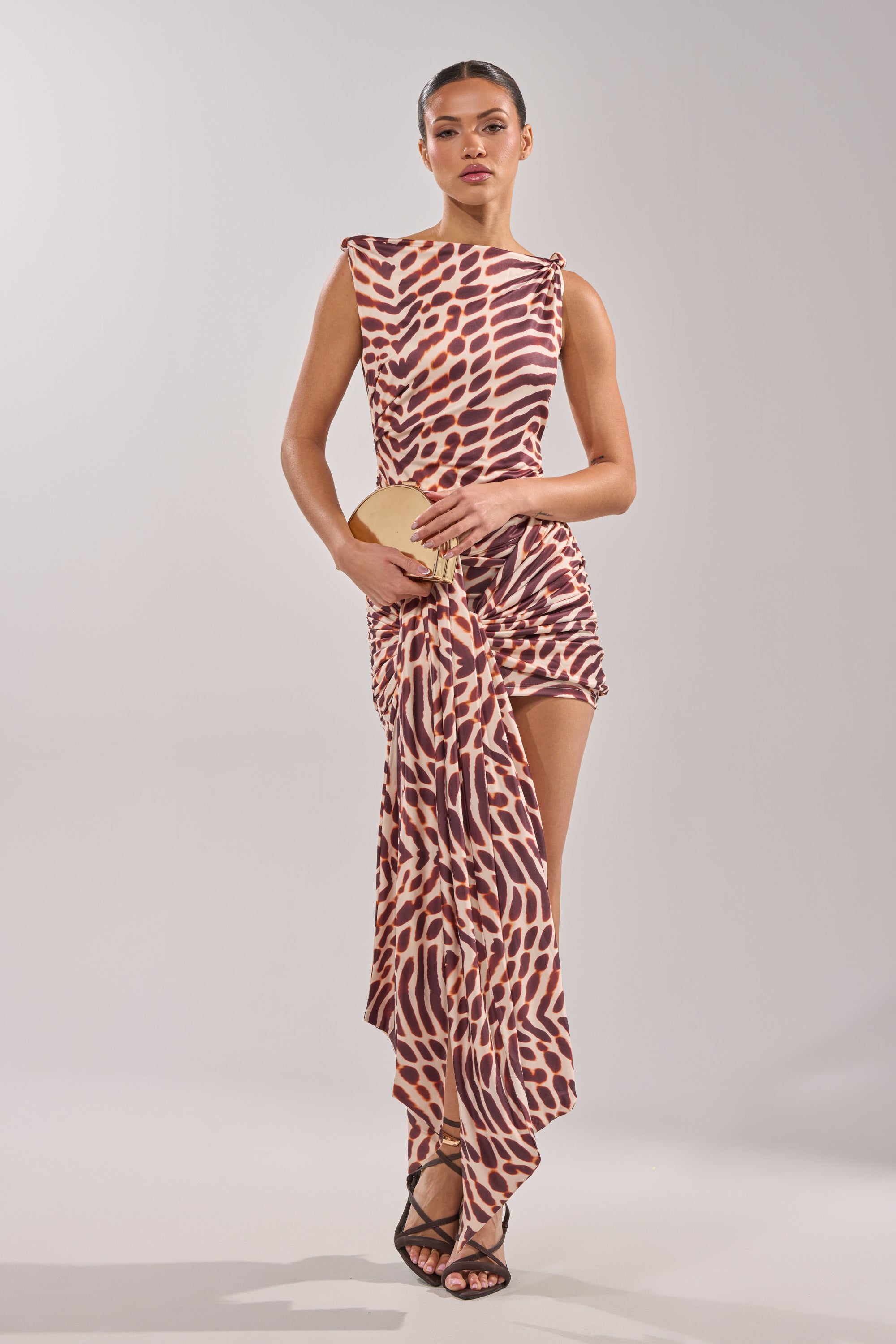 A woman poses against a plain background in the JUNGLE TIES LEOPARD MINI DRESS, an asymmetrical, brown and beige spotted mini dress with a draped skirt detail. She pairs it with black strappy heels and holds a circular beige clutch. 4