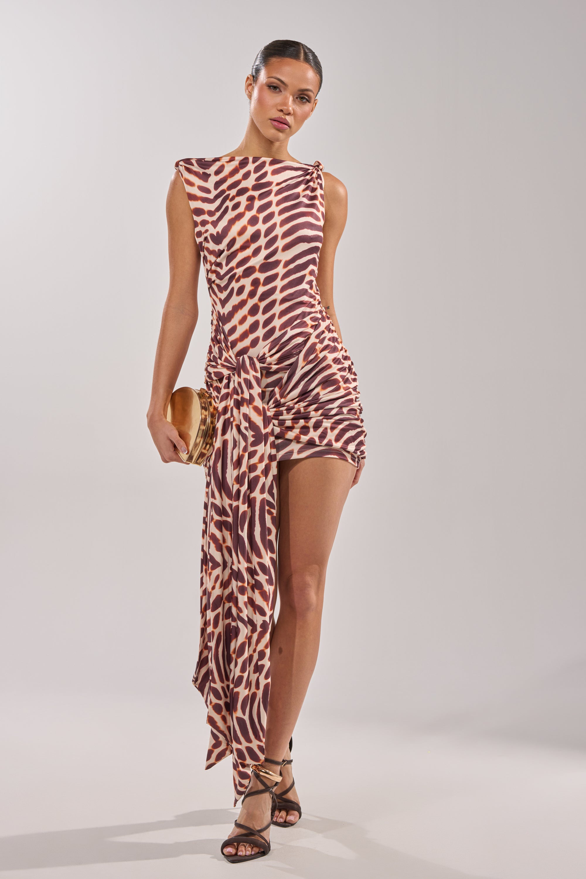 A woman stands against a plain light background wearing the JUNGLE TIES LEOPARD MINI DRESS, a sleeveless, asymmetrical mini dress with a brown and cream animal print, paired with strappy black heels and holding a clutch. 5