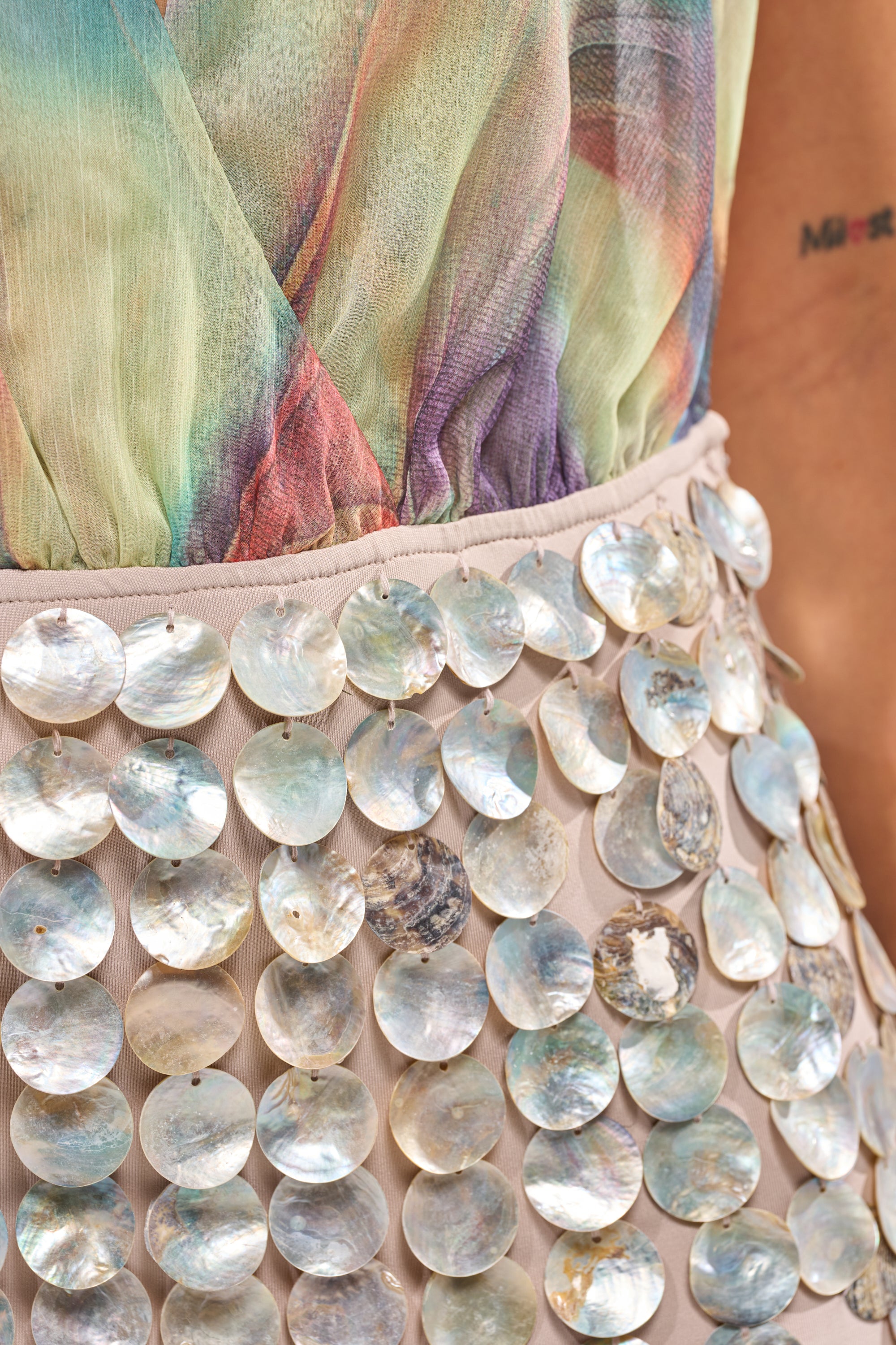 Close-up of a person wearing the I SEA PURPLE MINI DRESS, a pastel multicolored sheer piece with large iridescent circular sequins. An abstract resort look, with part of their arm and a small tattoo visible on the right. 8