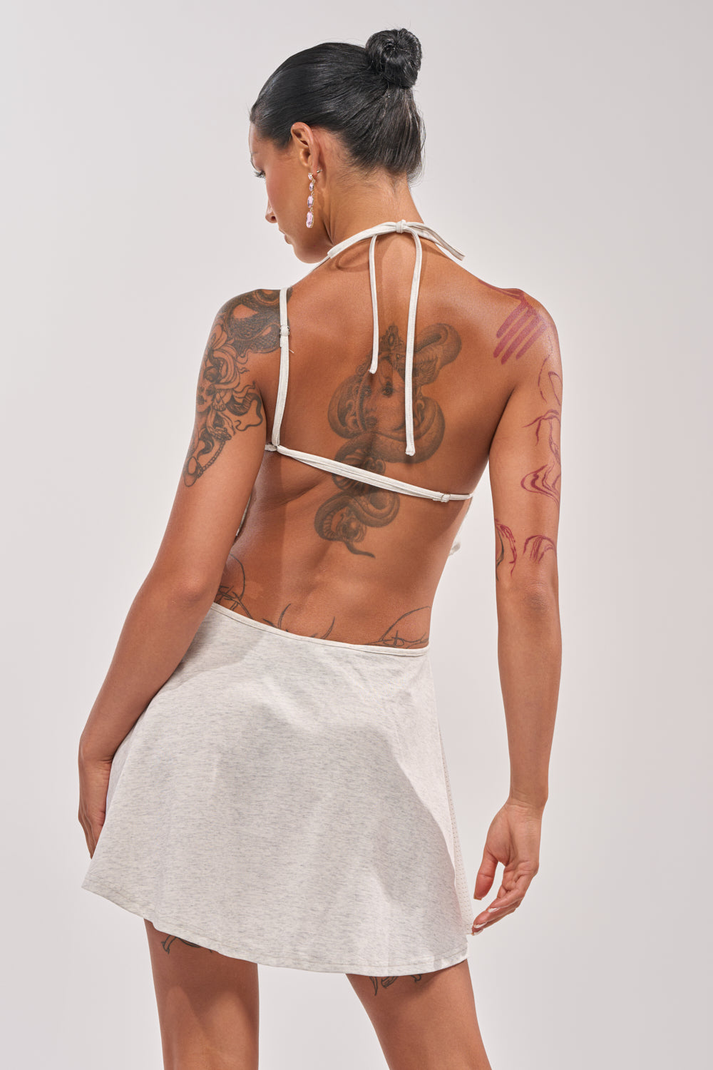 A woman with black hair in a bun, wearing the BETTER THAN THE NEXT MINI DRESS, stands with her back to the camera. She has visible tattoos and earrings, shown against a plain light background. 4