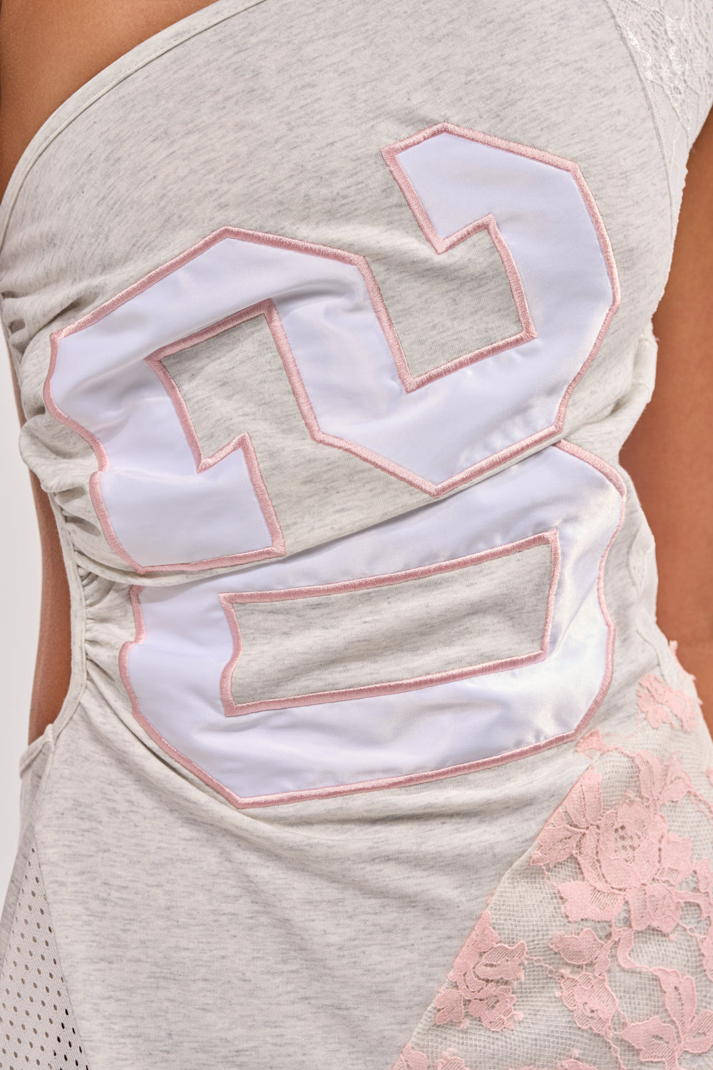 Close-up of a person wearing the BETTER THAN THE NEXT MINI DRESS, a light gray mini dress with an asymmetrical neckline, large white and pink “23” patch, light pink lace paneling, mesh accents, and gathered stitching. 7