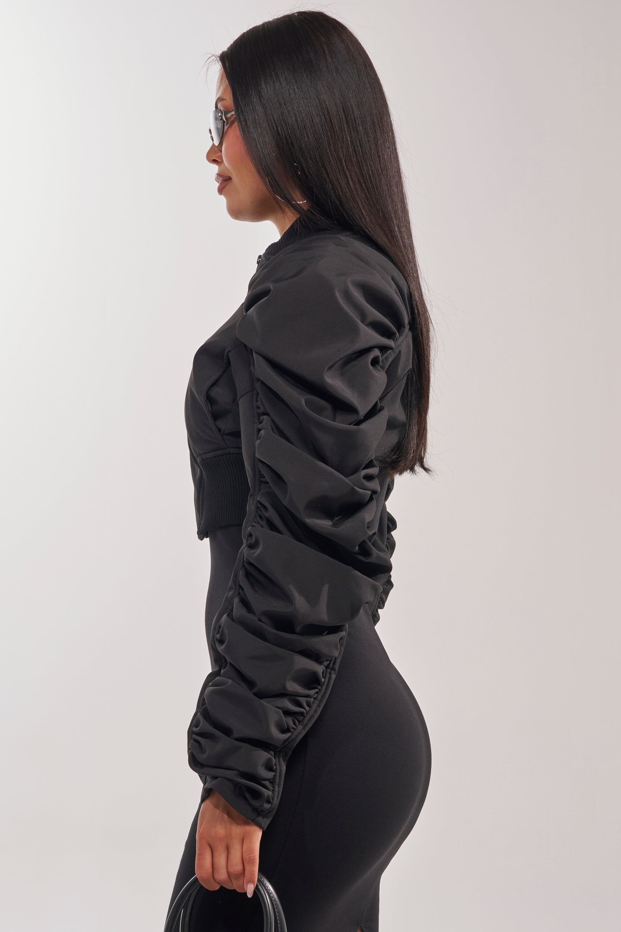 A woman with long straight hair and glasses stands in profile against a plain light background, wearing the NOTHING TO IT CROPPED BOMBER, black fitted pants, and holding a small black handbag. 5