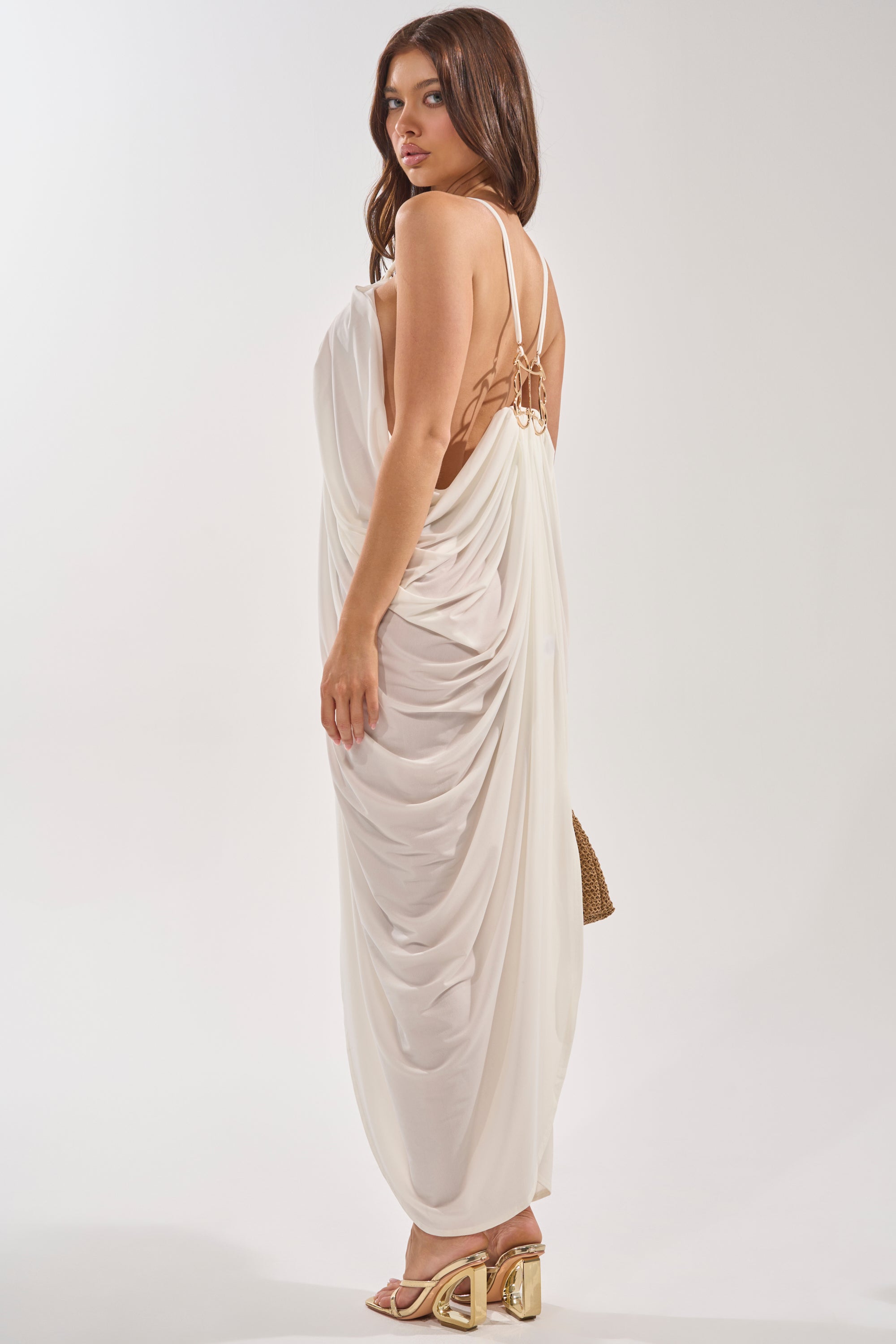A woman poses against a white background wearing the BALI MAMI LOW BACK MAXI DRESS IN WHITE with thin straps, paired with gold block-heeled sandals and a small purse. 0
