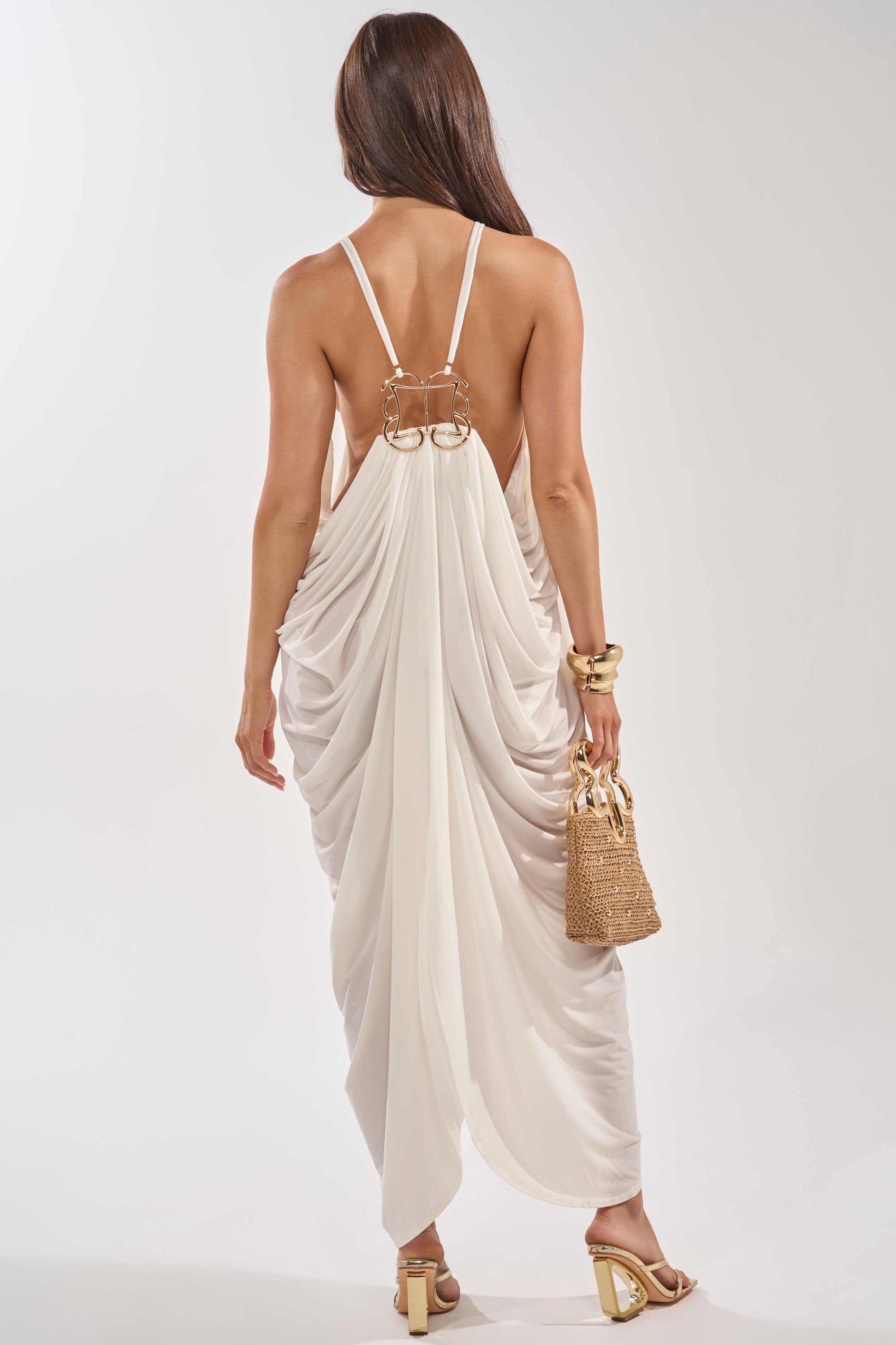A woman with long brown hair faces away in the BALI MAMI LOW BACK MAXI DRESS IN WHITE, styled with gold heels, bracelets, and a woven bag, capturing effortless resort style against a light background. 4
