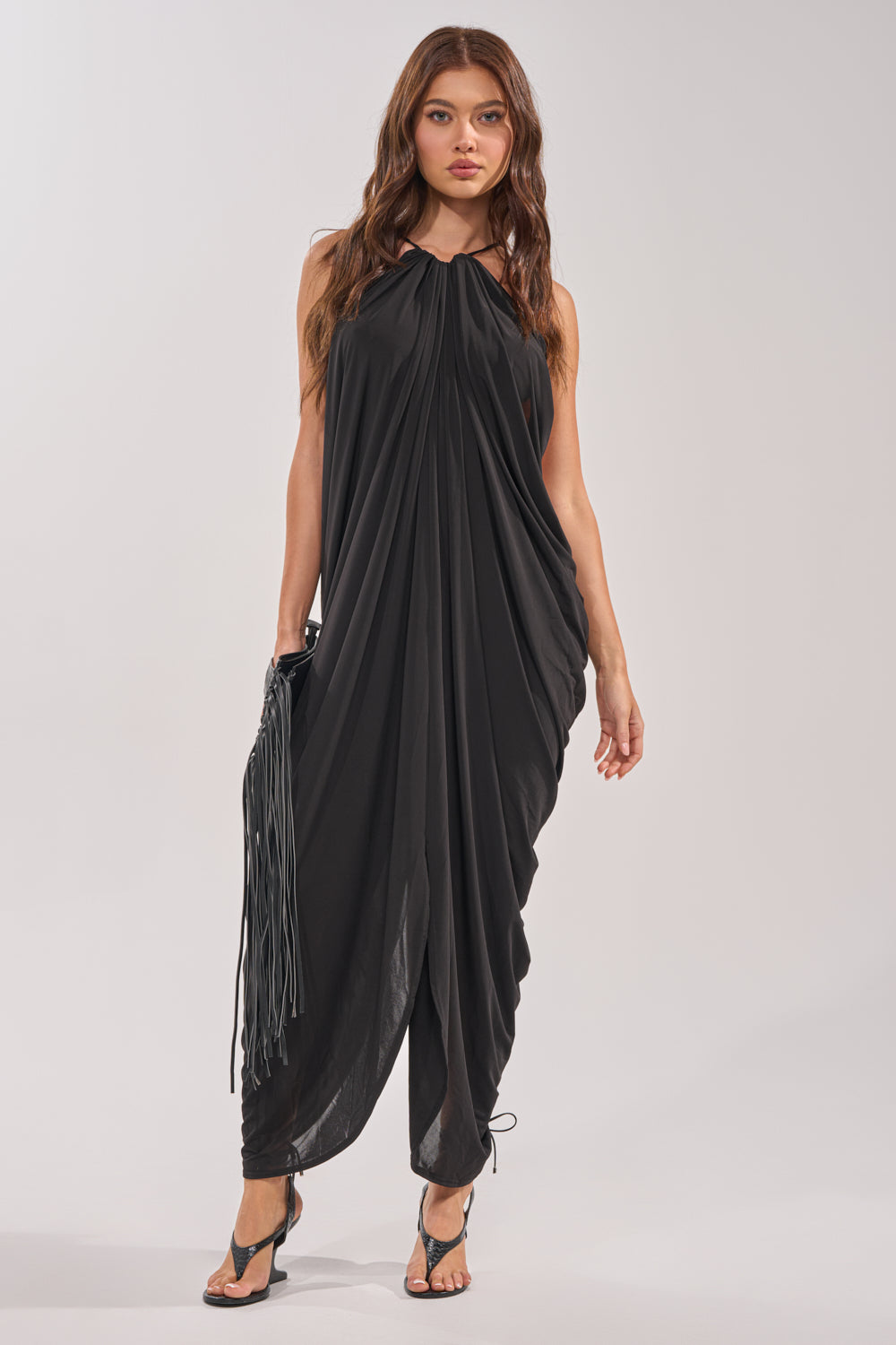 A woman poses against a plain background in the BALI MAMI LOW BACK MAXI DRESS IN BLACK, featuring a loose drape, gathered neckline, and low-cut back. She styles it with black heeled sandals and a fringed bag—ideal for any occasion. 1