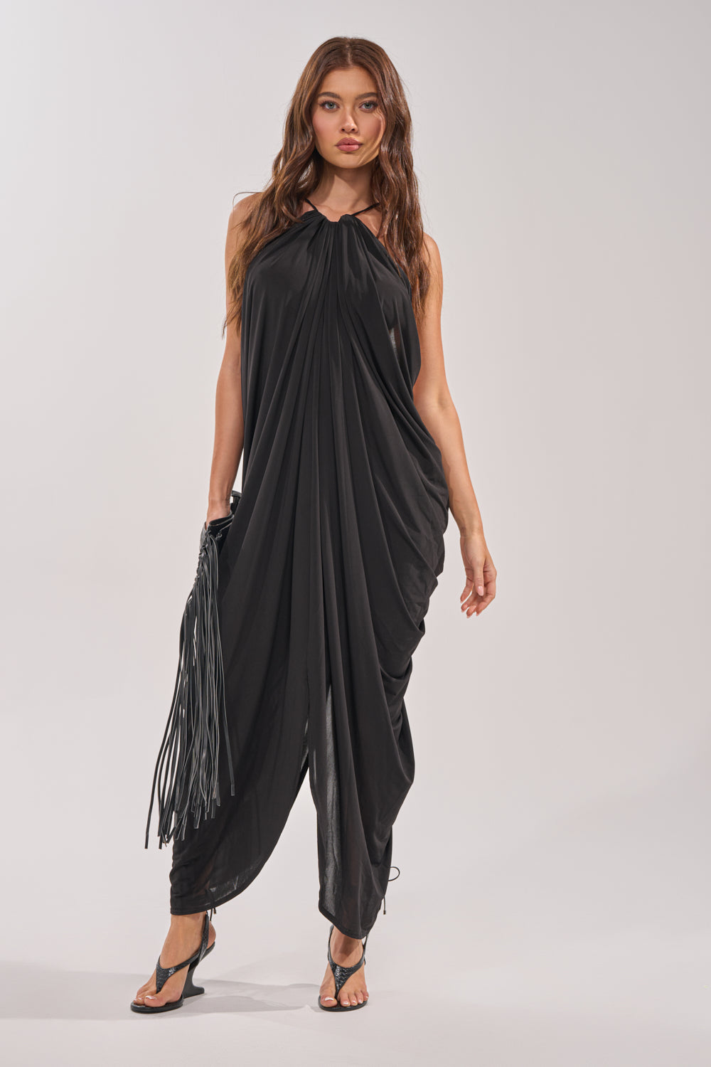 A woman wears the BALI MAMI LOW BACK MAXI DRESS IN BLACK, styled for special occasions. She holds a black fringed bag and pairs the dress with black heeled sandals against a plain light background. 2