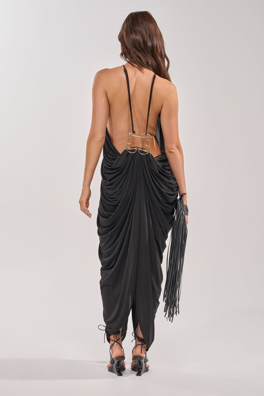 A woman with long brown hair is shown from the back, wearing the BALI MAMI LOW BACK MAXI DRESS IN BLACK with gold clasps and strappy heels. She carries a black fringed handbag, standing out against a light background. 4