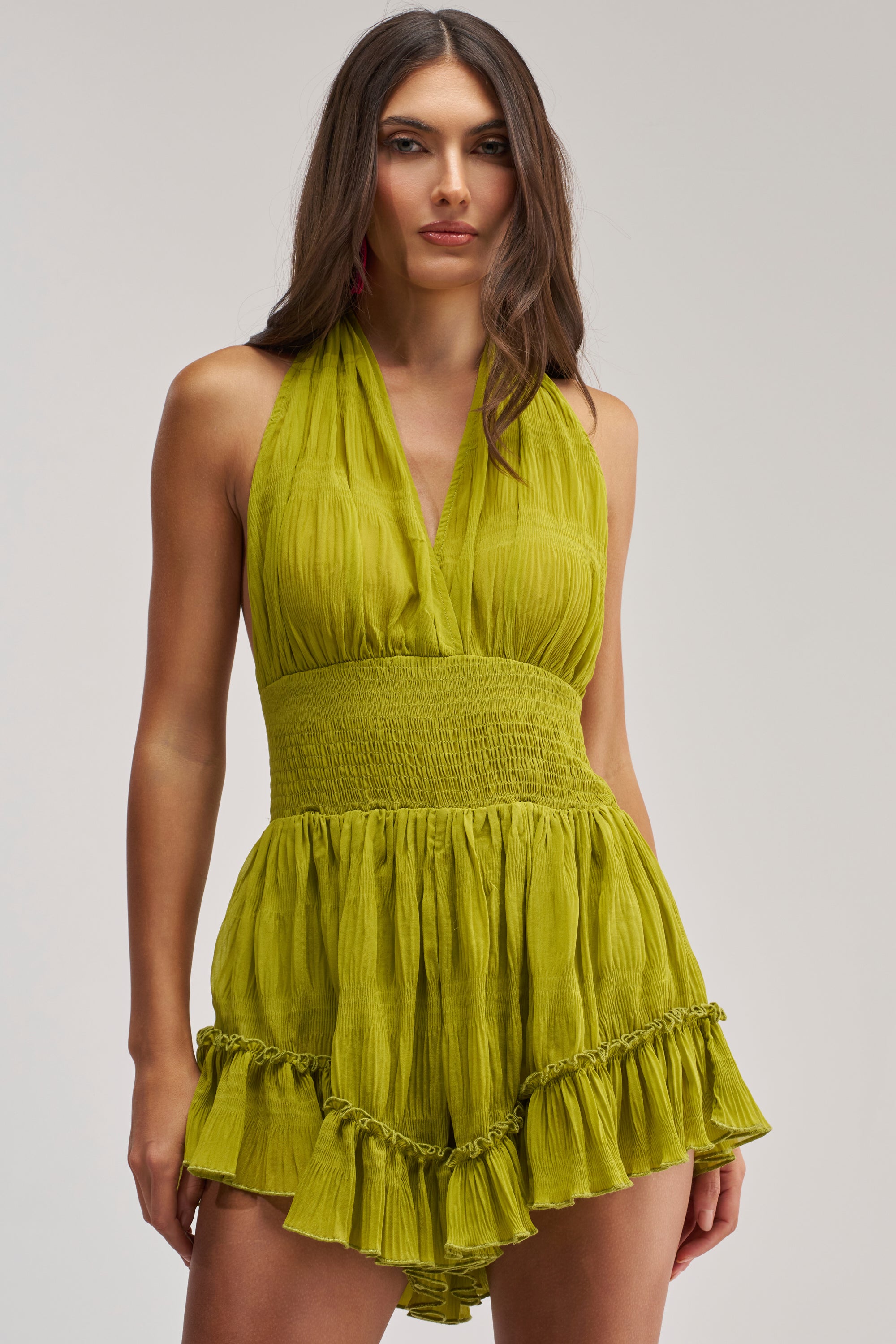 A woman with long brown hair models the SUMMER DAY CHIFFON SLEEVELESS ROMPER in lime green, styled with a halter-neck, ruched waist, and ruffled hem—an effortless vacation look against a plain background.