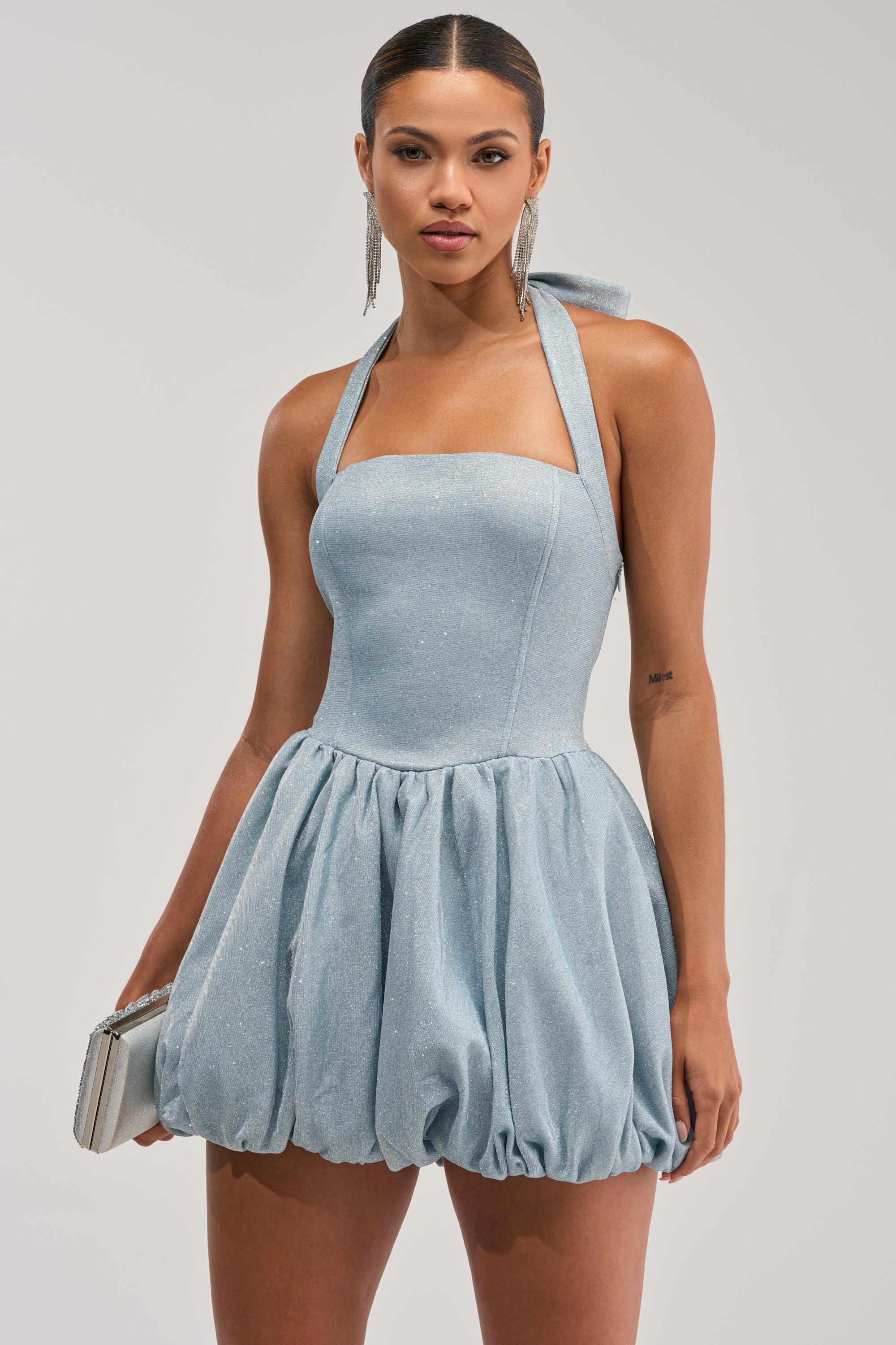 A woman stands against a plain background in the GLISTEN UP SHIMMER HALTER MINI DRESS, a light blue sparkly mini dress with a bubble skirt. She holds a silver clutch, wears long earrings, and has sleek hair with a confident look.