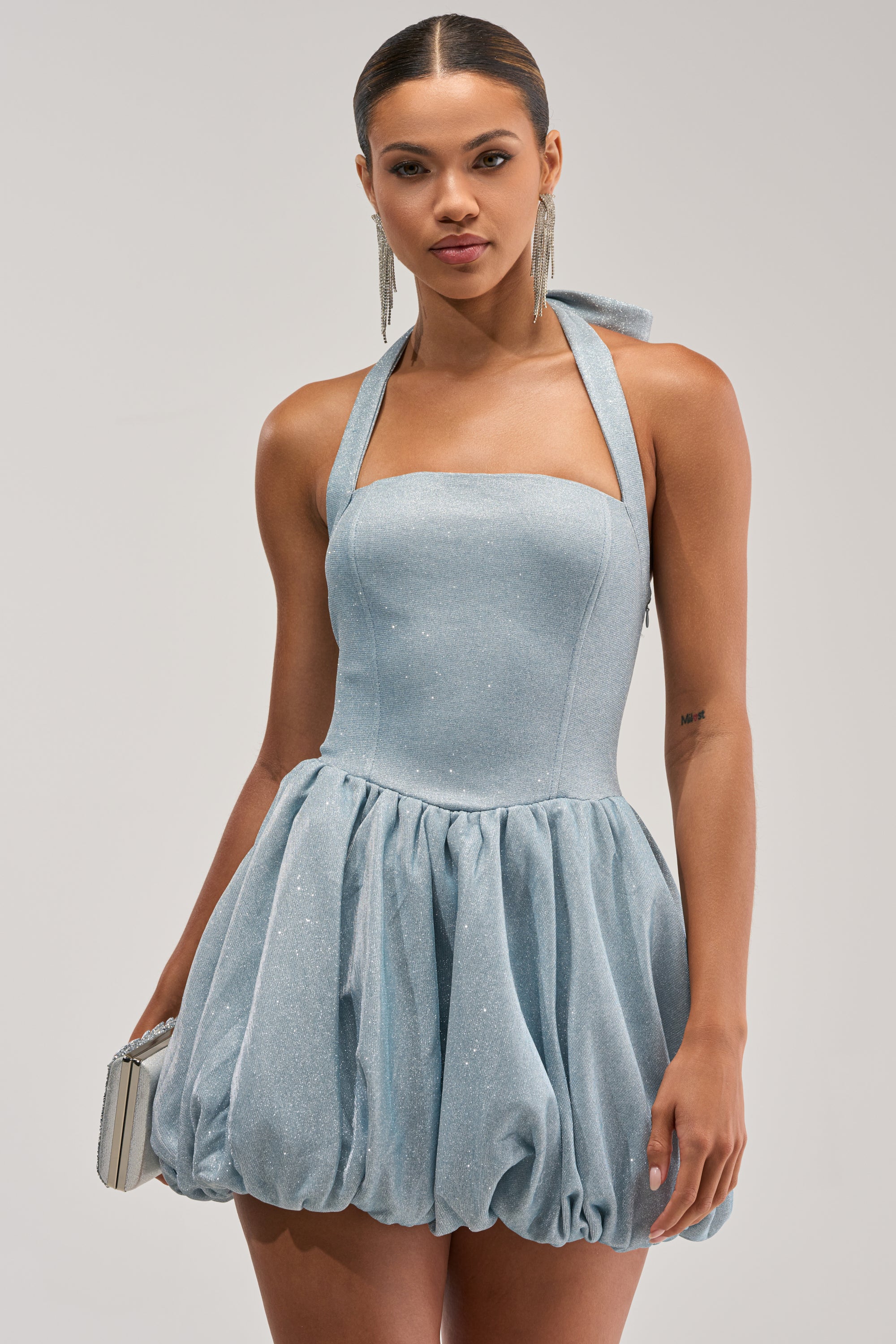 A woman in the GLISTEN UP SHIMMER HALTER MINI DRESS—sparkly light blue with a fitted bodice and bubble skirt—holds a silver clutch and wears long earrings.