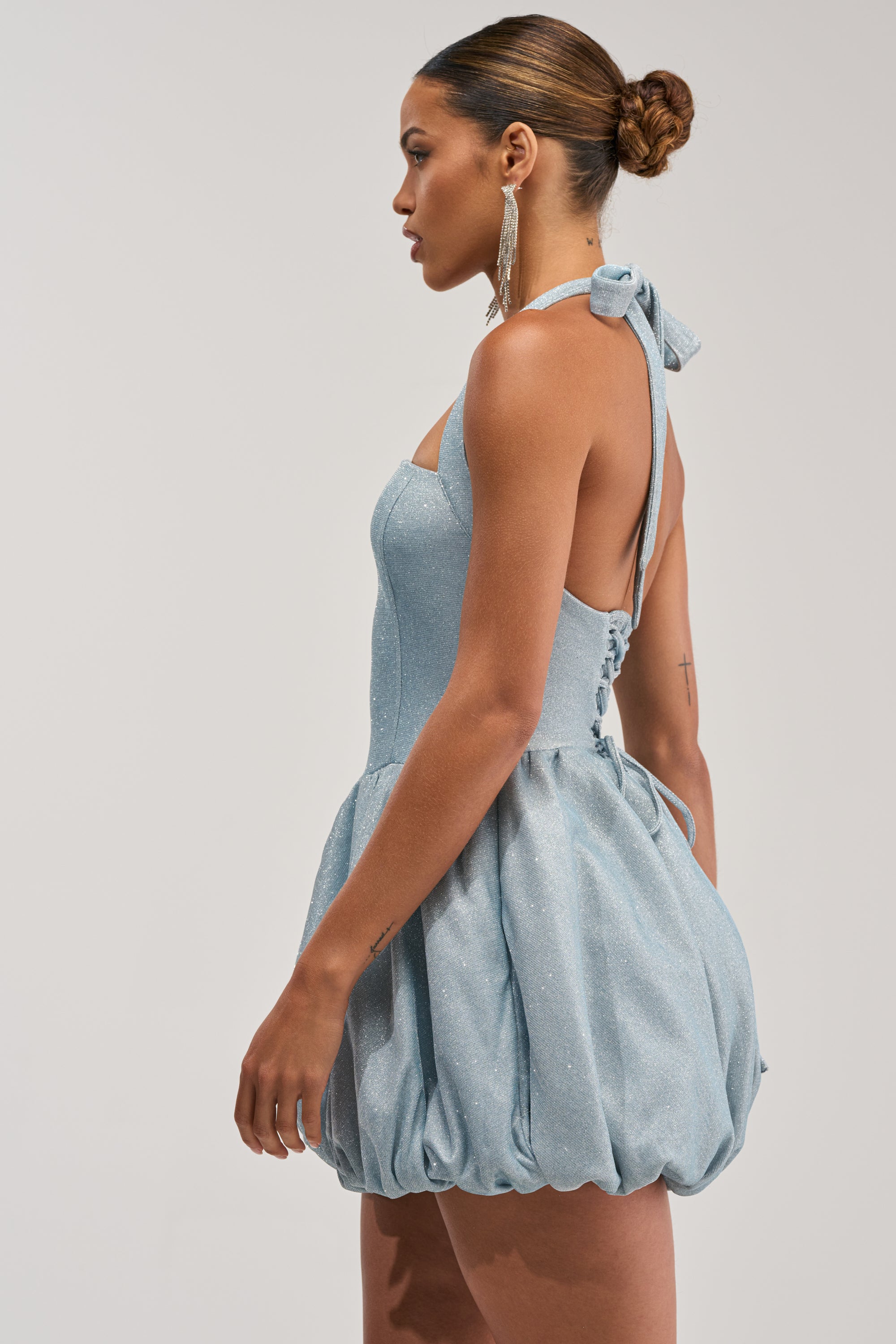 A woman in profile wears the GLISTEN UP SHIMMER HALTER MINI DRESS, a light blue sparkly halter-neck dress with a fitted bodice and bubble skirt. Her hair is in a neat bun, paired with long dangling earrings.
