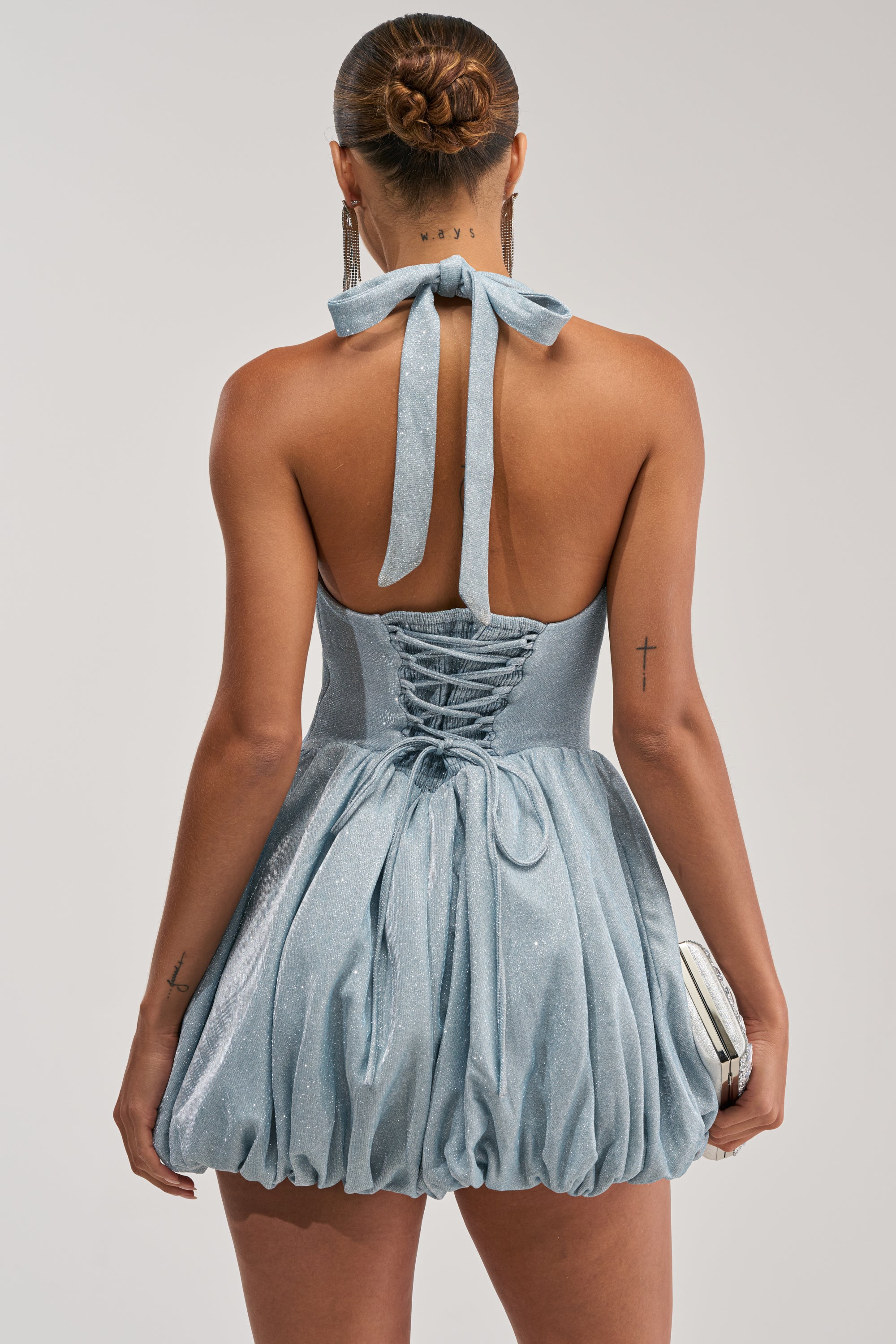 A woman is shown from the back wearing the GLISTEN UP SHIMMER HALTER MINI DRESS in light blue, featuring a bow and lace-up detail. She holds a silver clutch, her hair is in a neat bun, and visible tattoos are on her arms.