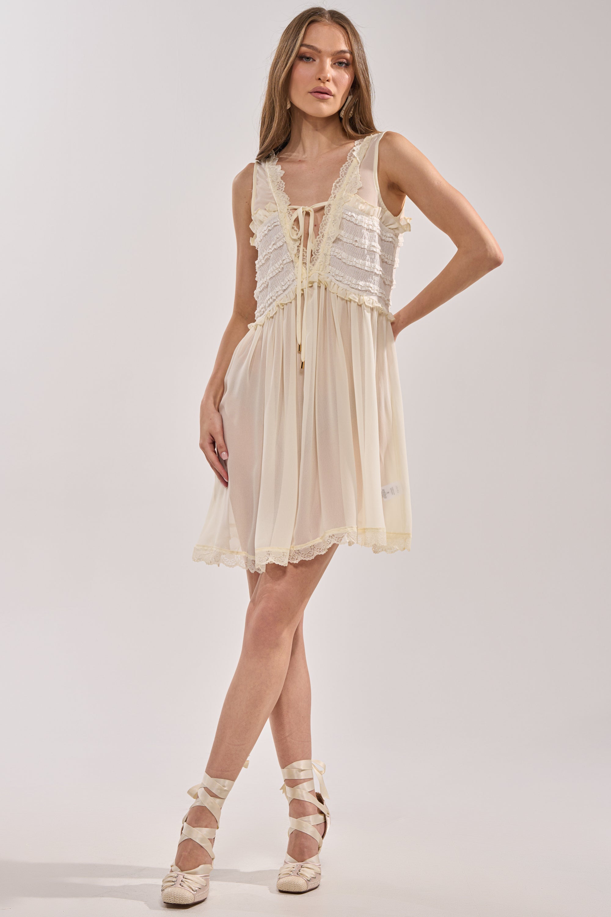 A woman poses against a light background wearing the FREE SPIRIT LACE MINI DRESS, an ivory, sleeveless mini dress with ruffles and lace details finished with a bow at the neckline, paired with ballet-inspired lace-up shoes. 0