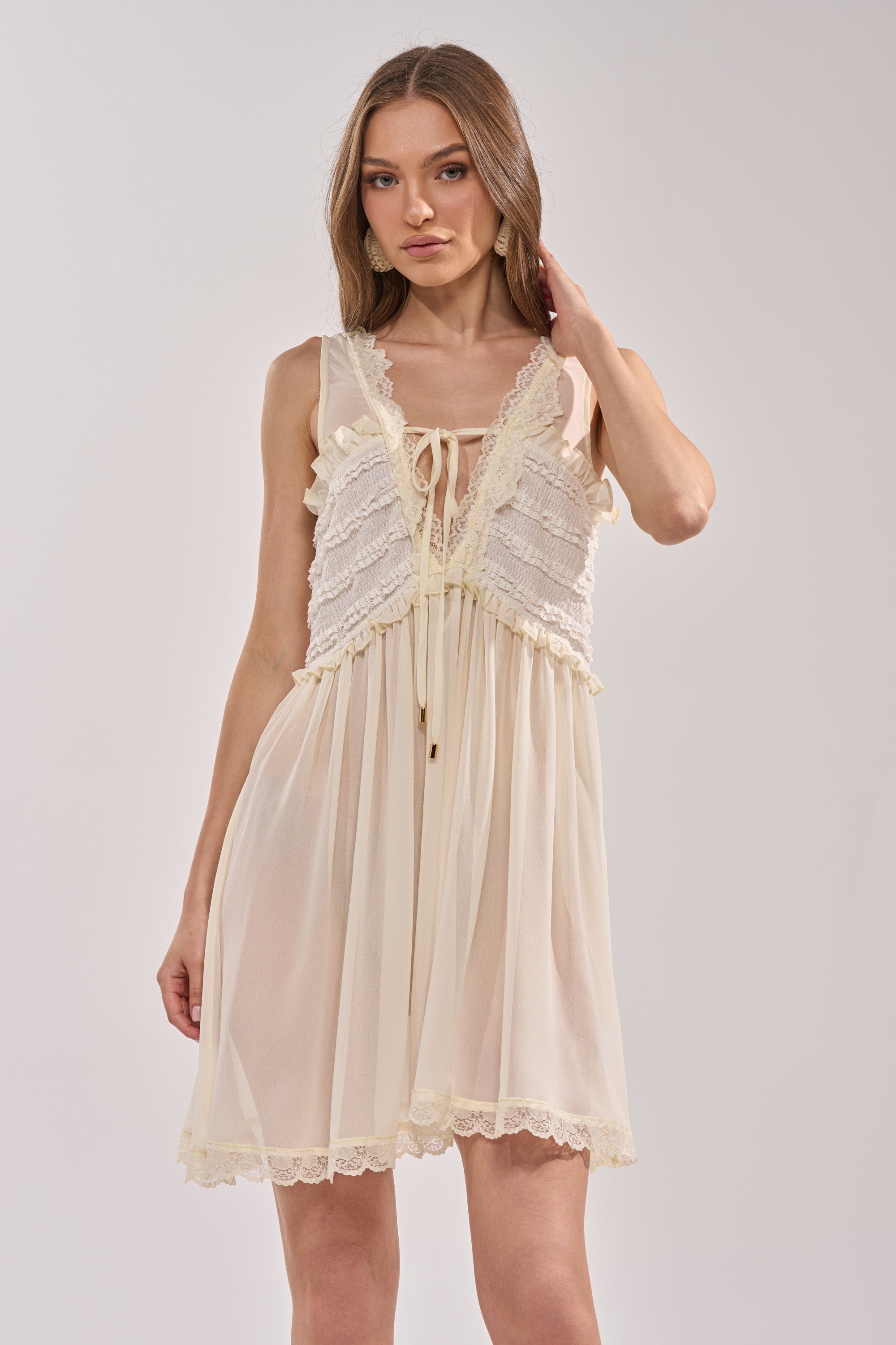A woman in the FREE SPIRIT LACE MINI DRESS—ivory with a ruffled lace bodice and sheer skirt—stands against a plain background, one hand in her hair; a perfect choice for summer brunch. 1