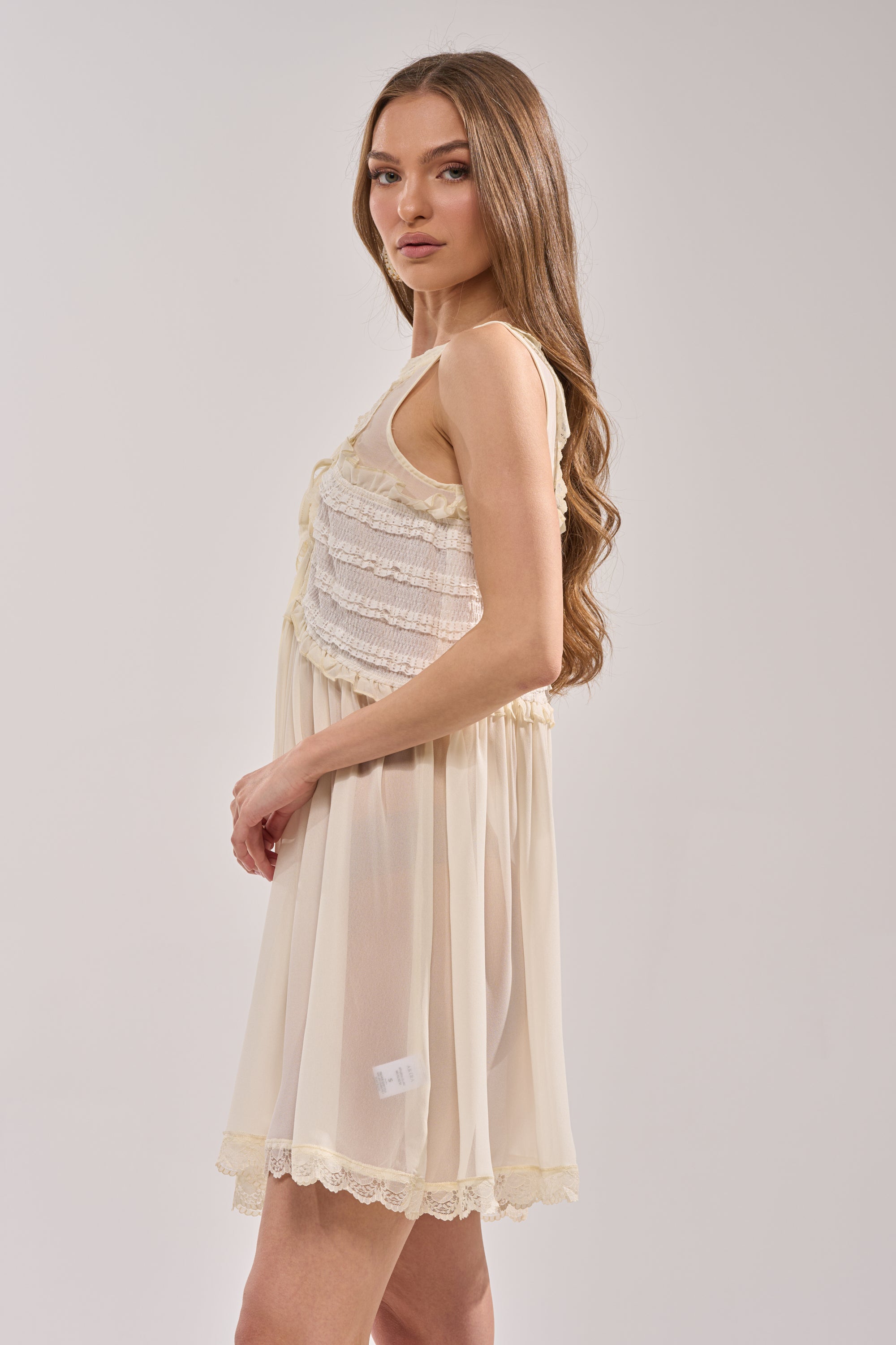 A young woman with long, wavy brown hair models the FREE SPIRIT LACE MINI DRESS in ivory with ruffle lace trim. She stands sideways against a plain light background, creating an effortless summer brunch look. 2