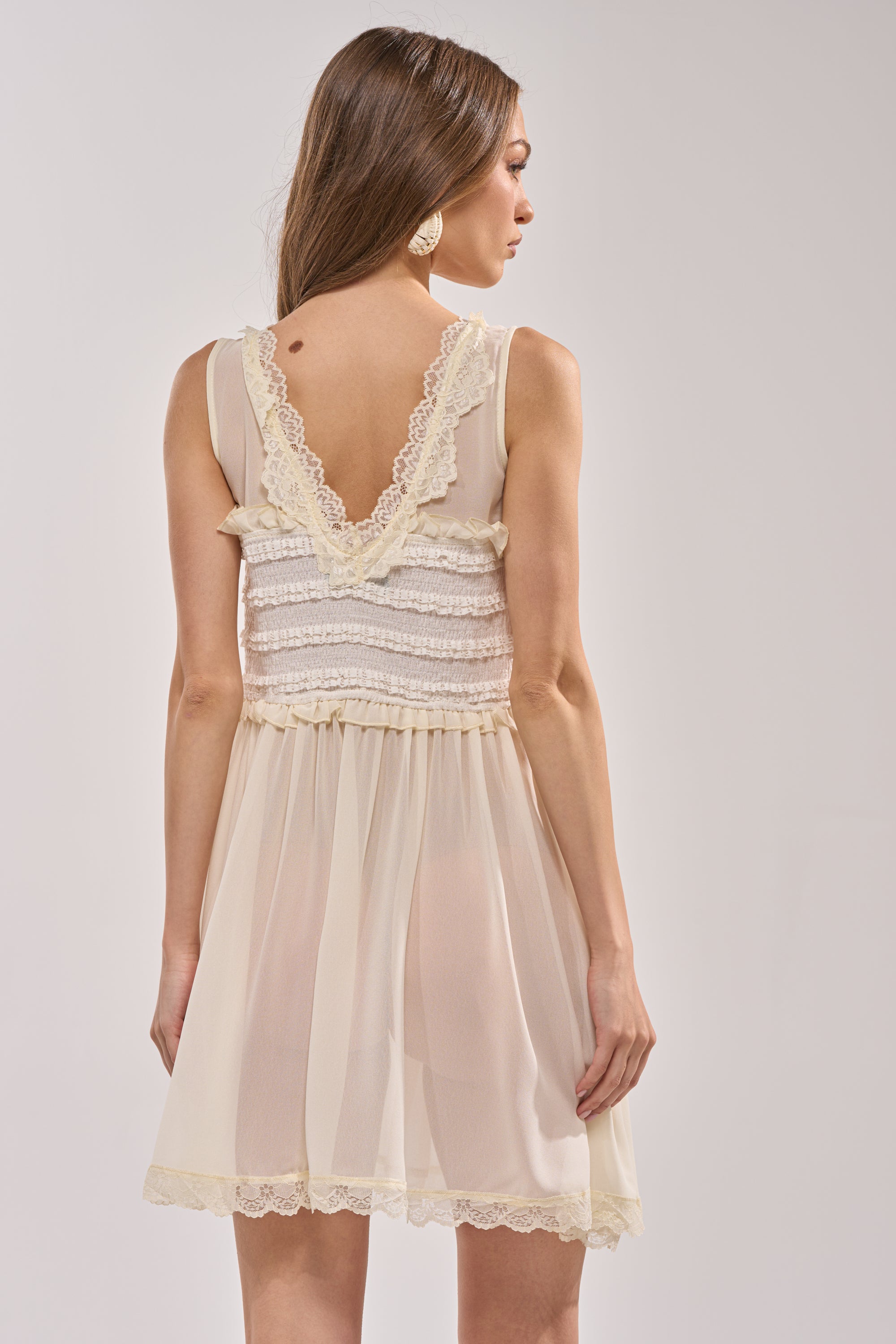 A woman faces away from the camera in the FREE SPIRIT LACE MINI DRESS, a sleeveless ivory mini with lace-trimmed ruffles and a V-shaped ruffled back, showcasing effortless boho-chic summer style. 3