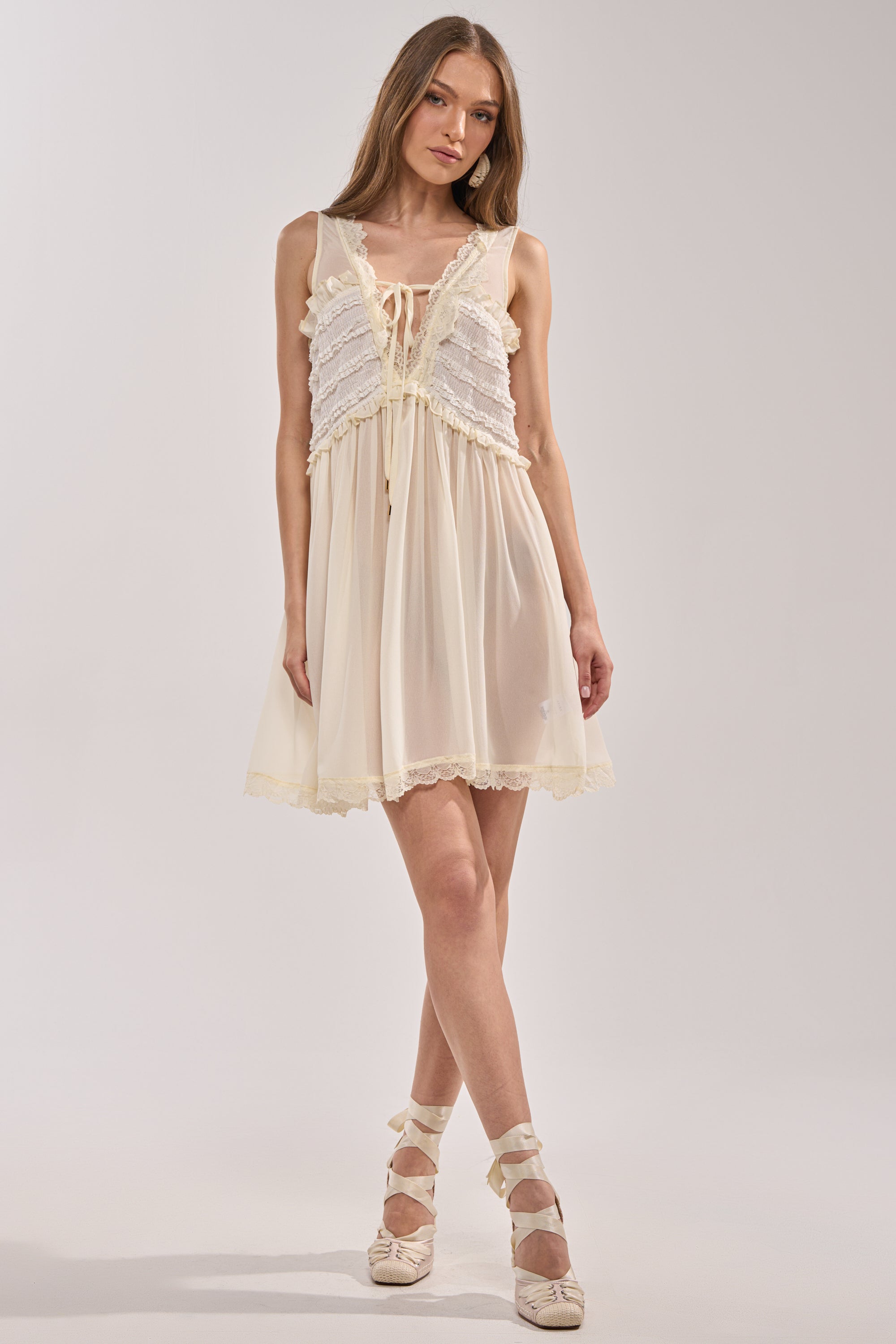 A woman wears the FREE SPIRIT LACE MINI DRESS in ivory with ruffle details and lace trim, standing on one leg in matching lace-up ballet flats against a plain light background. 4