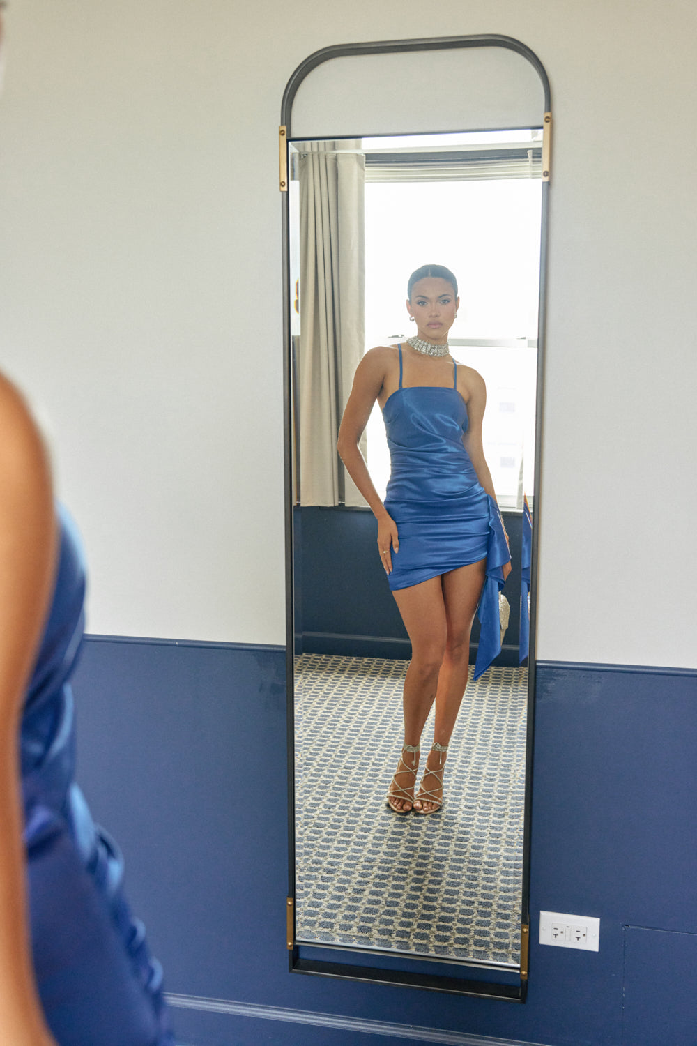 Wearing the TWIRL ME TWICE SATIN MINI DRESS and heels, a woman stands before a tall mirror, admiring her reflection in a blue and white room illuminated by light streaming through the window behind her.