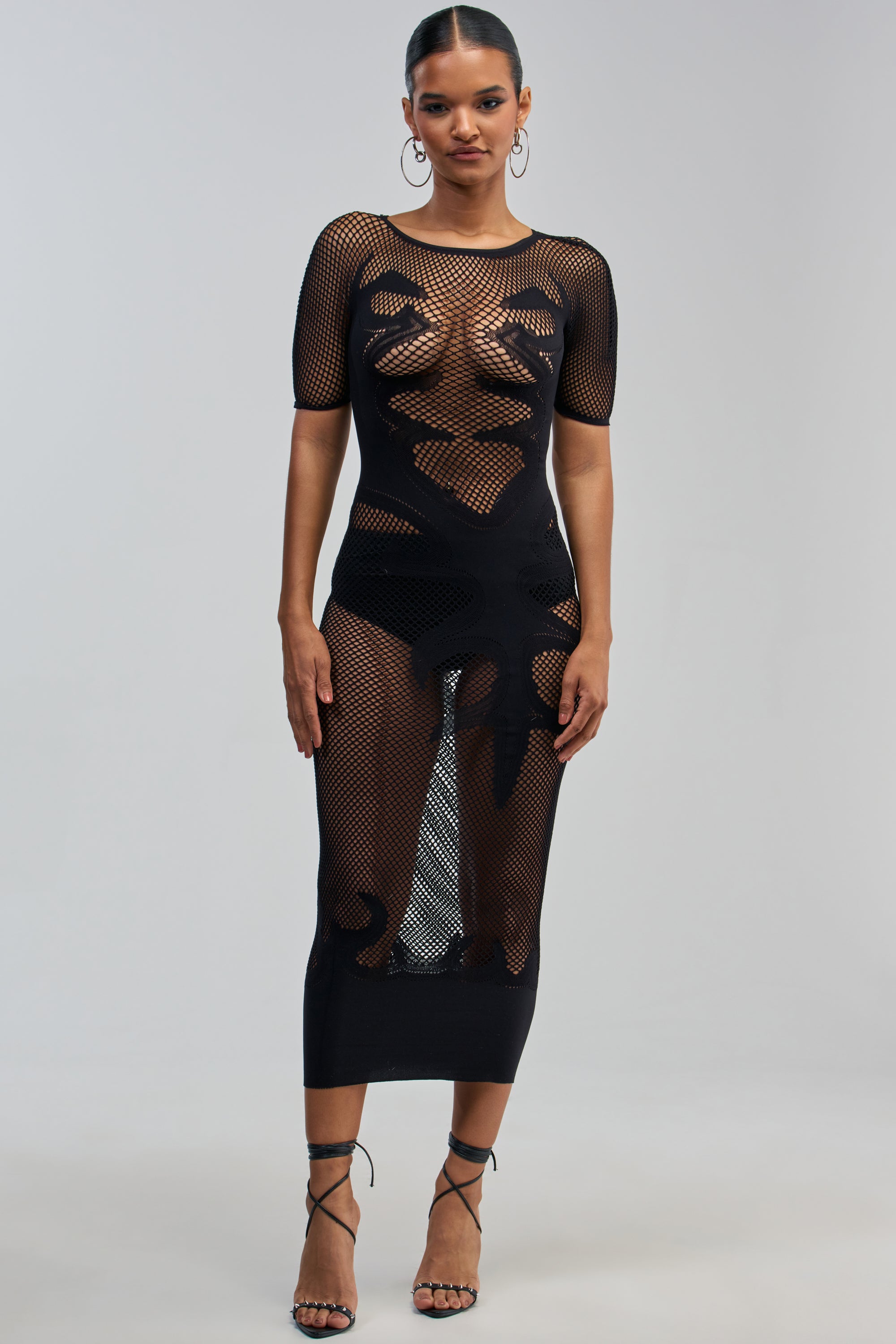 A woman stands facing forward, wearing the CLUB HOPPER SUPER STRETCH BODYCON MAXI IN BLACK—a sexy night-out dress featuring bold abstract patterns on sheer black mesh. Visible beneath is a black bikini-style undergarment. She accessorizes with hoop earrings, strappy black heels, and has her hair tied back.