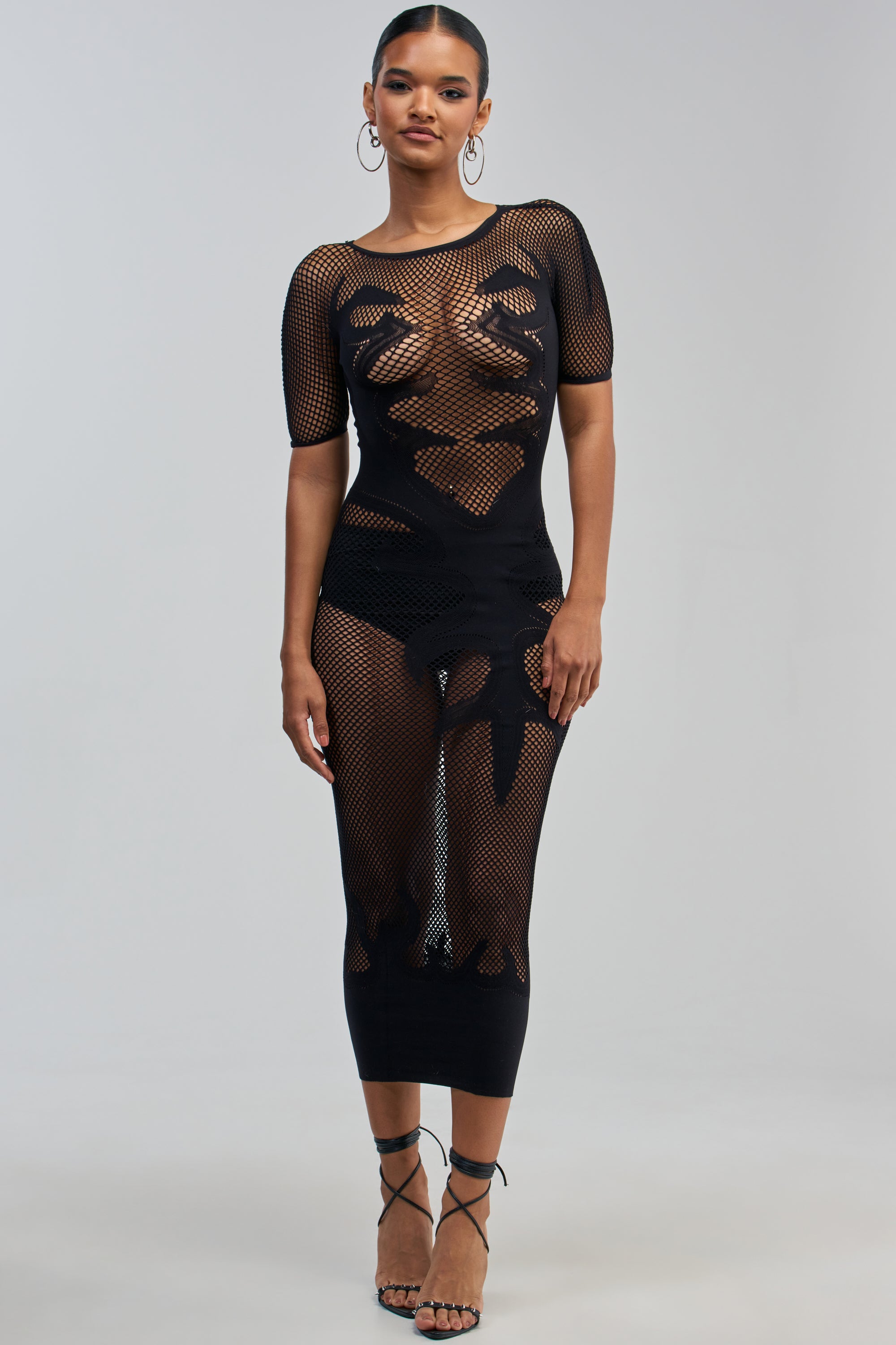 A woman stands facing forward, wearing the CLUB HOPPER SUPER STRETCH BODYCON MAXI IN BLACK—a sheer black mesh dress with abstract patterns evoking the allure of a sexy night out. The outfit features short sleeves and strappy heels, perfectly capturing the appeal of this form-fitting bodycon maxi dress. The background is plain and light-colored.