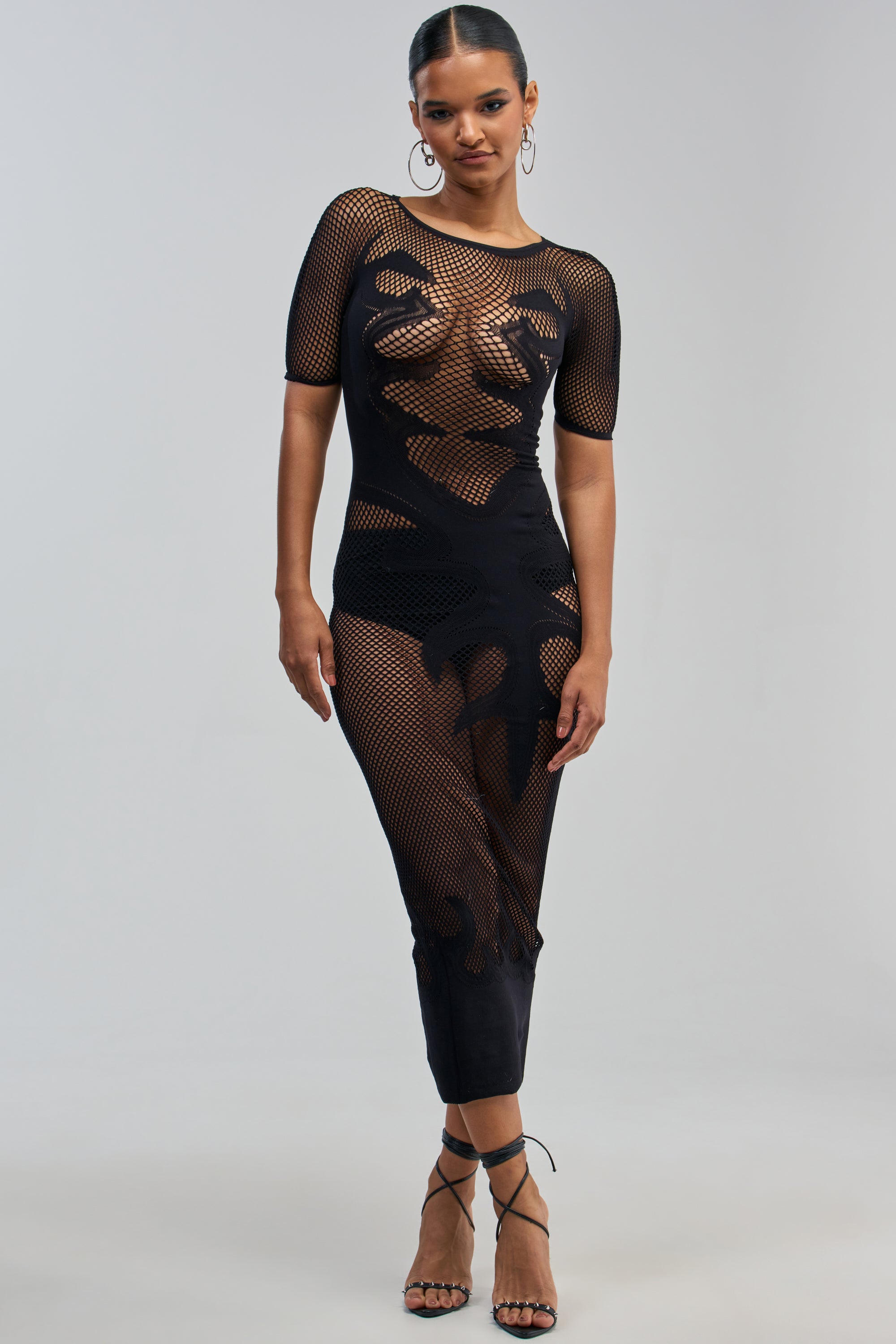 A woman stands confidently in the CLUB HOPPER SUPER STRETCH BODYCON MAXI IN BLACK, a black short-sleeved mesh bodycon maxi dress featuring abstract opaque patterns. She pairs it with strappy black high heels and hoop earrings, her hair pulled back as she poses against a plain, light background.