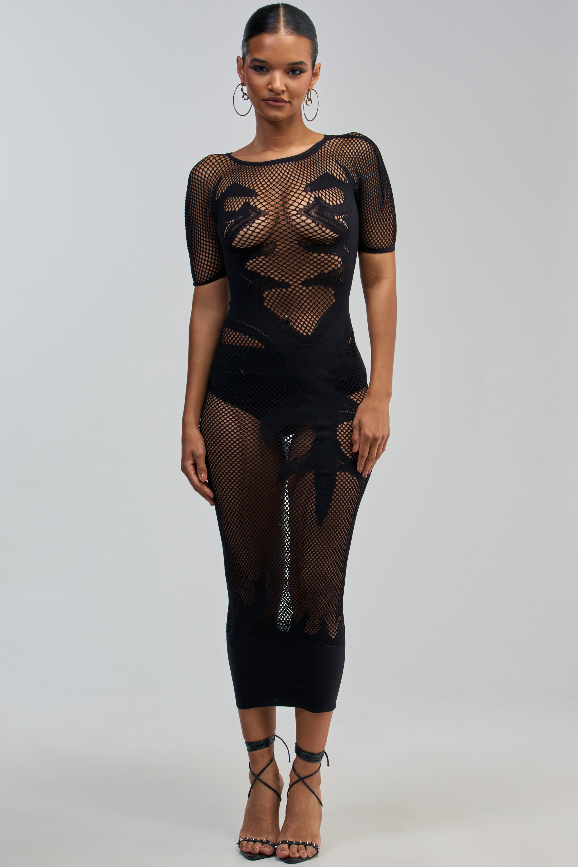 Facing forward, a woman wears the CLUB HOPPER SUPER STRETCH BODYCON MAXI IN BLACK—a sheer black mesh dress with abstract patterns, short sleeves, and a fitted silhouette—paired with strappy high heels against a plain light gray background.