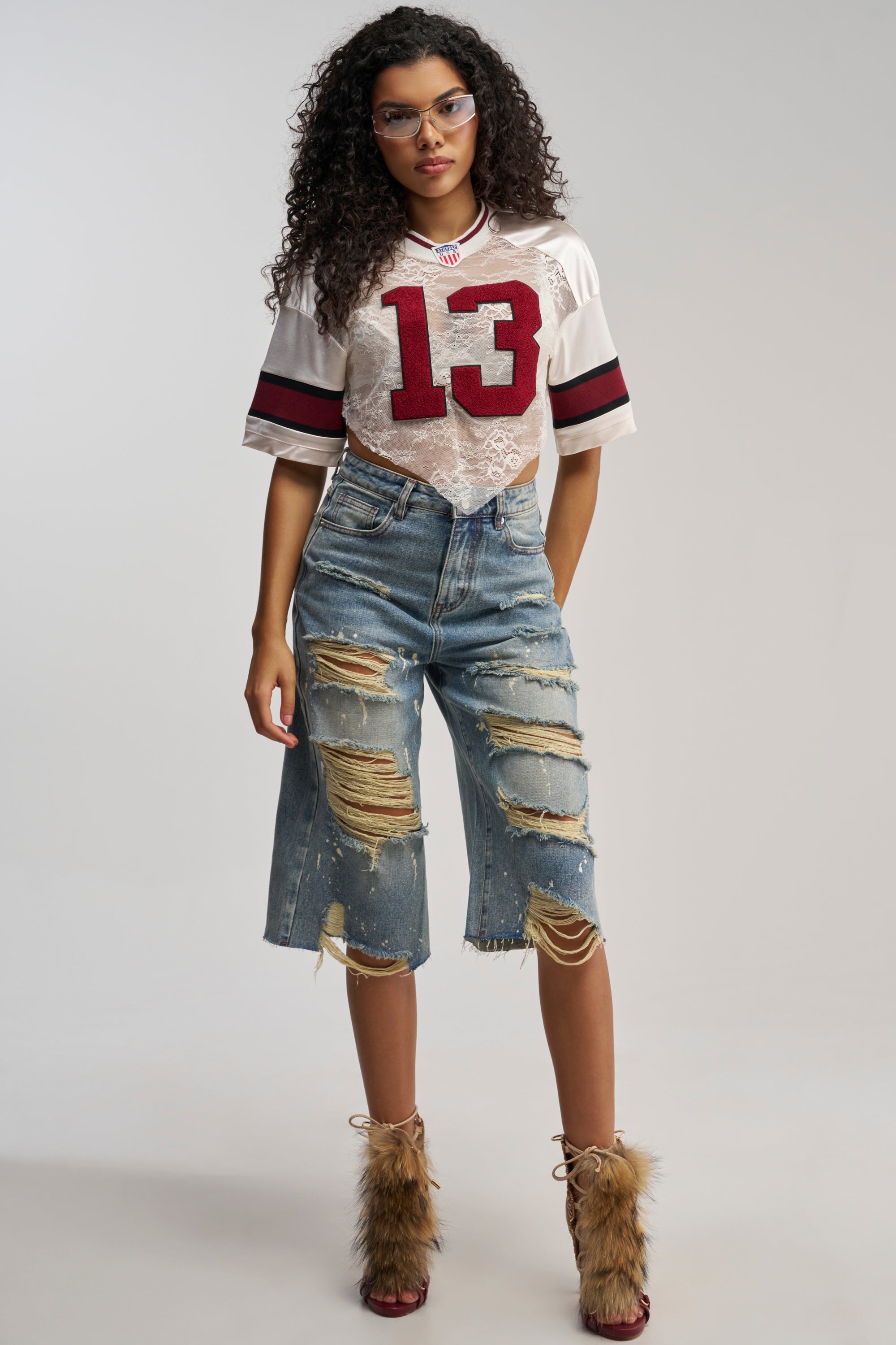 A woman with curly hair and glasses exudes casual streetwear style as she stands confidently against a plain background, sporting a white crop jersey with the number 13, fur-trimmed heels, and the FEELING DISTRESSED BERMUDA SHORT in a medium wash.