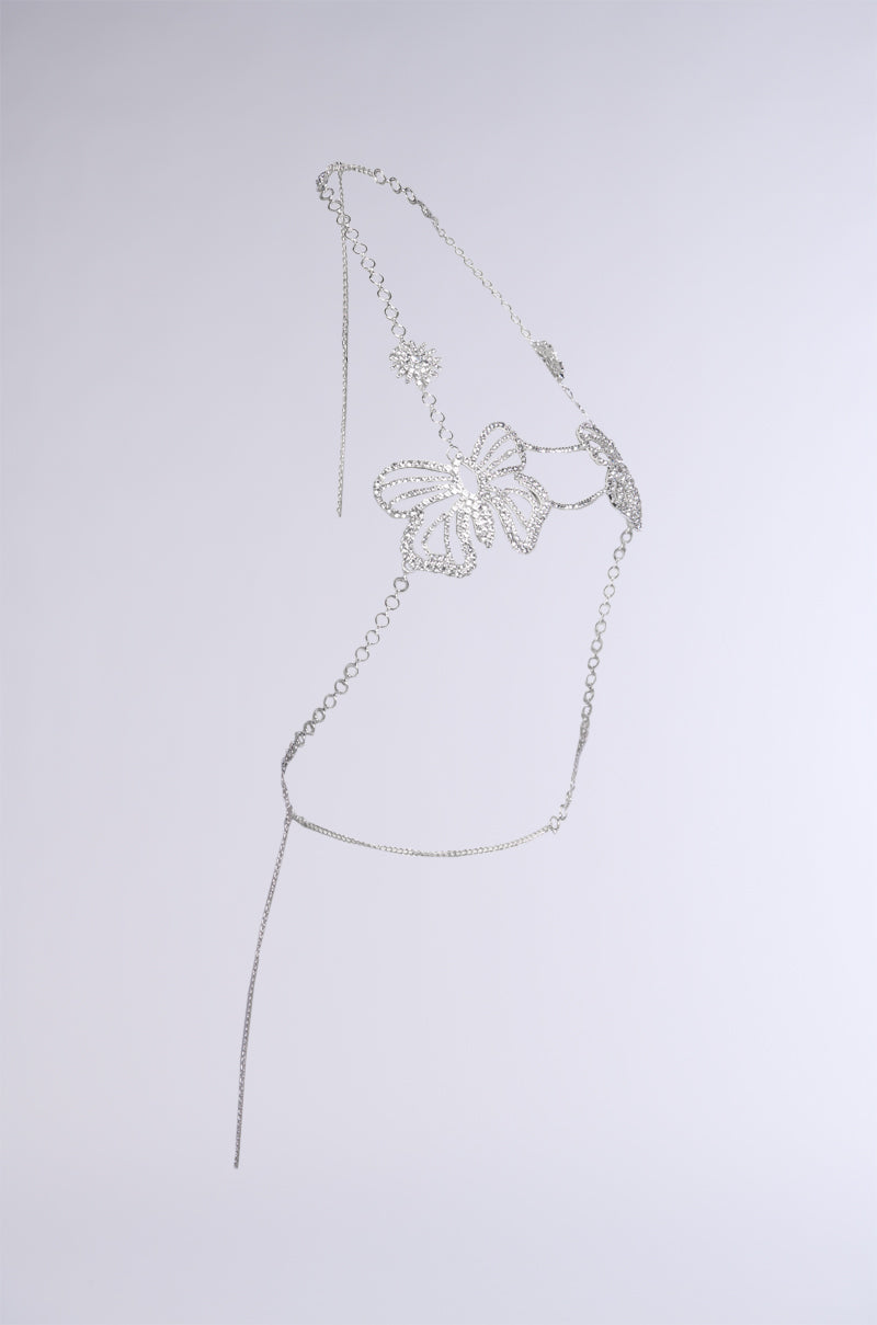 A delicate and intricate silver metal sculpture, titled "WATCH ME FLY BODY CHAIN," is suspended against a plain white background. The artwork features a large butterfly design at its center, evocative of the body chain's central motif, with circular link chains extending from the ends to create a light and airy appearance.