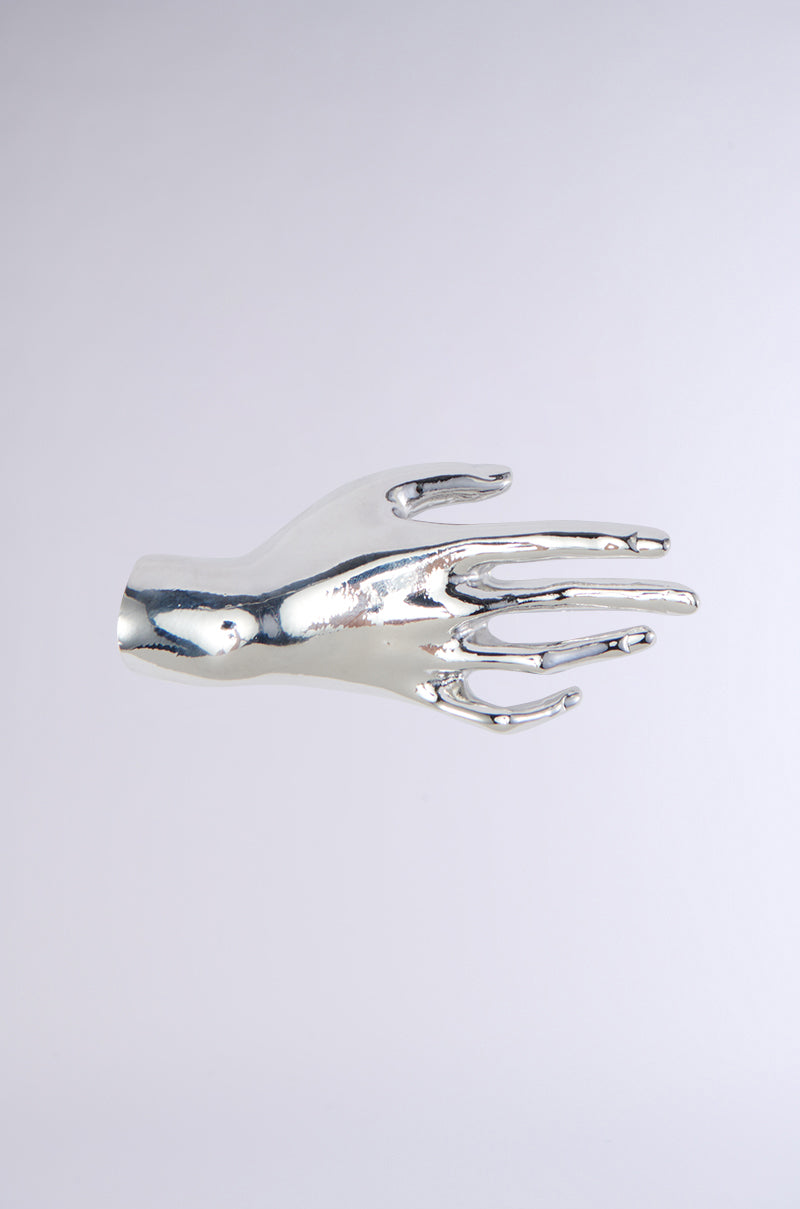 A polished faux silver metallic sculpture, named HANDS ON BROOCH, is shown against a plain, light background. The brooch showcases a smooth and reflective right hand with fingers slightly bent and close together, making it a striking statement accessory.
