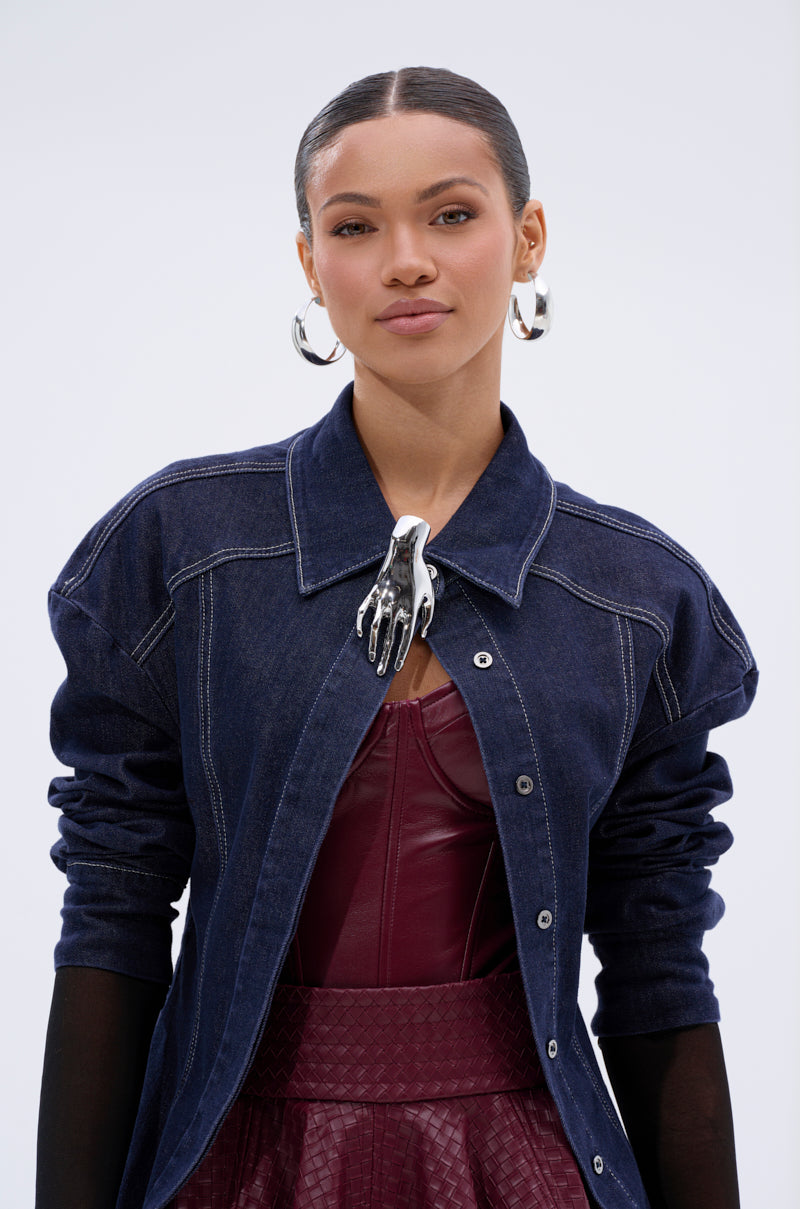 A woman with slicked-back hair wears large faux silver hoop earrings, a dark denim jacket, a burgundy leather top, and a skirt. The HANDS ON BROOCH clips to her jacket collar as a striking statement accessory against the light background.