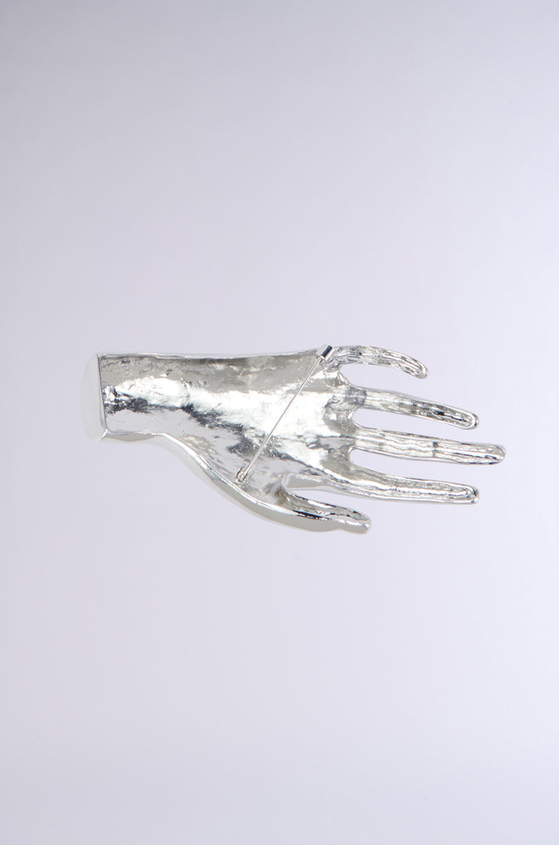 The HANDS ON BROOCH is a faux silver metallic sculpture representing a right hand that's slightly curved and open, featuring intricately detailed fingers. Set against a light gray background, the shiny surface truly stands out. This statement accessory exudes a minimalist and modern tone, making it the perfect hands-on brooch.