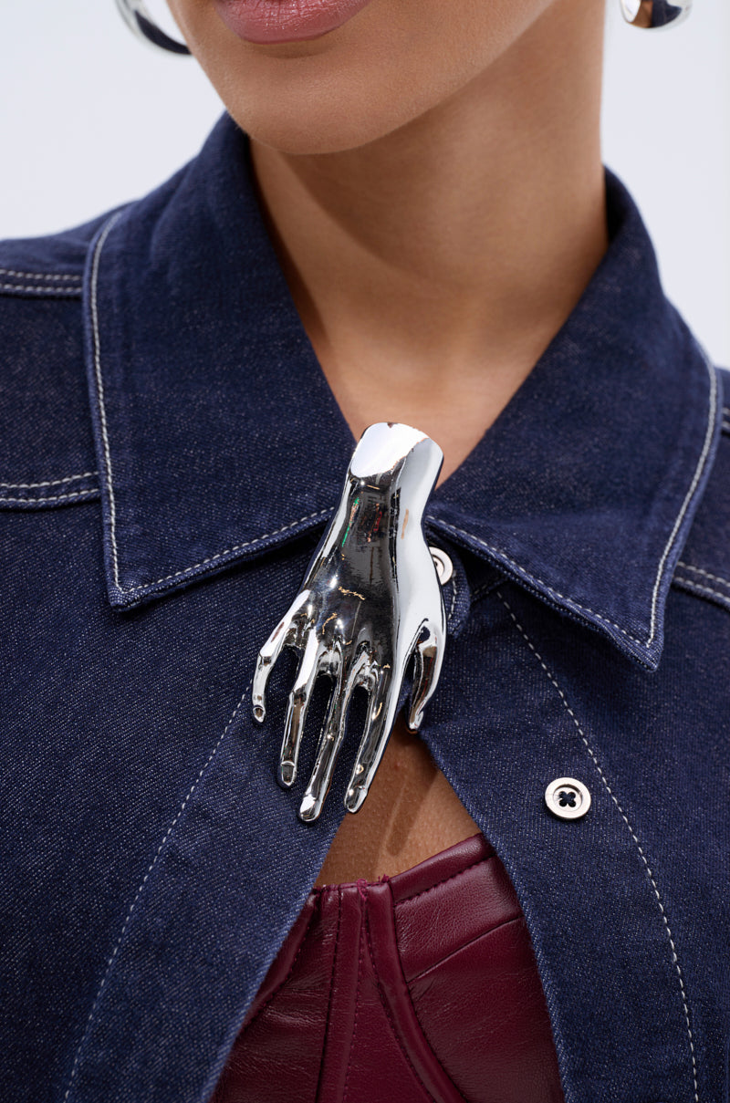 A close-up shows a person in a dark denim jacket with white stitching, a burgundy leather top, and the HANDS ON BROOCH—a bold faux silver accessory—clipped to their collar. Part of the face and earrings are also visible.