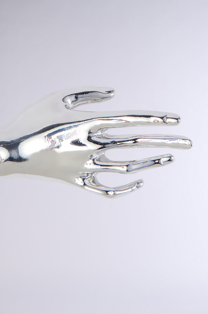 A close-up of the HANDS ON BROOCH, with its sleek, shiny metallic finish and reflective surface. The outstretched hand design is set against a plain, light-colored background. Its elongated and smooth fingers create a futuristic, abstract appearance reminiscent of an avant-garde faux silver metallic statement accessory.