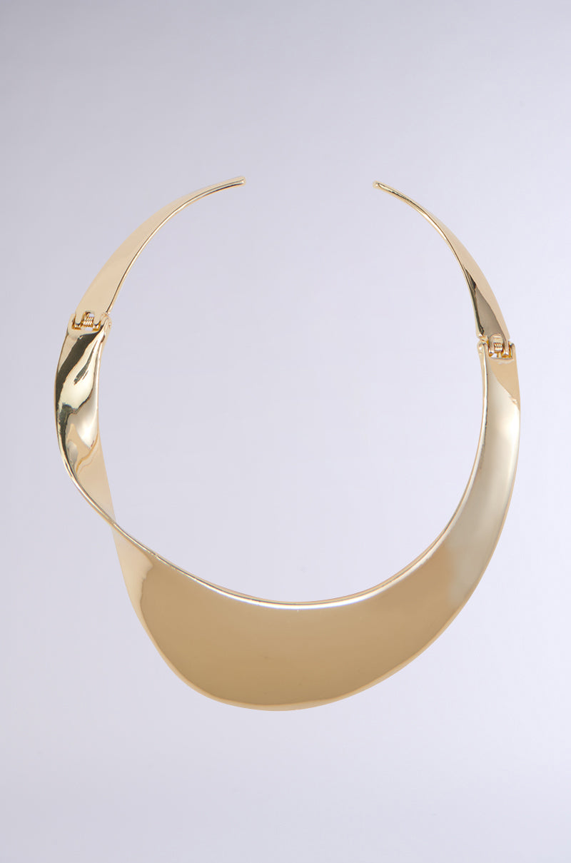 The TOP TIER CHOKER NECKLACE is a sleek, curved faux gold choker with a reflective surface. Its design showcases an asymmetrical, fluid shape that delicately twists around the neck. This piece of statement jewelry embodies a minimalistic and elegant appearance, creating a bold yet refined impression.