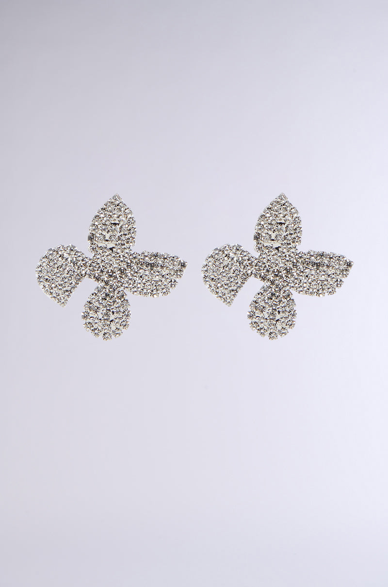 The SHOW ME LOVE EARRING is a pair of silver, diamond-studded earrings shaped like four-petaled flowers, set against a gray gradient background. They feature intricate detailing and sparkling silver rhinestones that reflect light, highlighting their elegance.