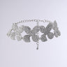 The SHOW ME LOVE CHOKER NECKLACE is a silver choker adorned with glittering, leaf-shaped embellishments. This statement piece features an intricate arrangement of crystal-studded leaves and an adjustable chain closure with a dangling charm for fitting, set against a neutral gray background that highlights its elegance.