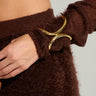 A close-up captures someone in a cozy, brown knitted outfit with their hand resting on their hip, displaying the striking EVERY OCCASION CUFF. This faux gold bracelet, featuring an elegant design of intertwined curves, stands out against the simple, neutral backdrop—ideal for cuffing season's style.