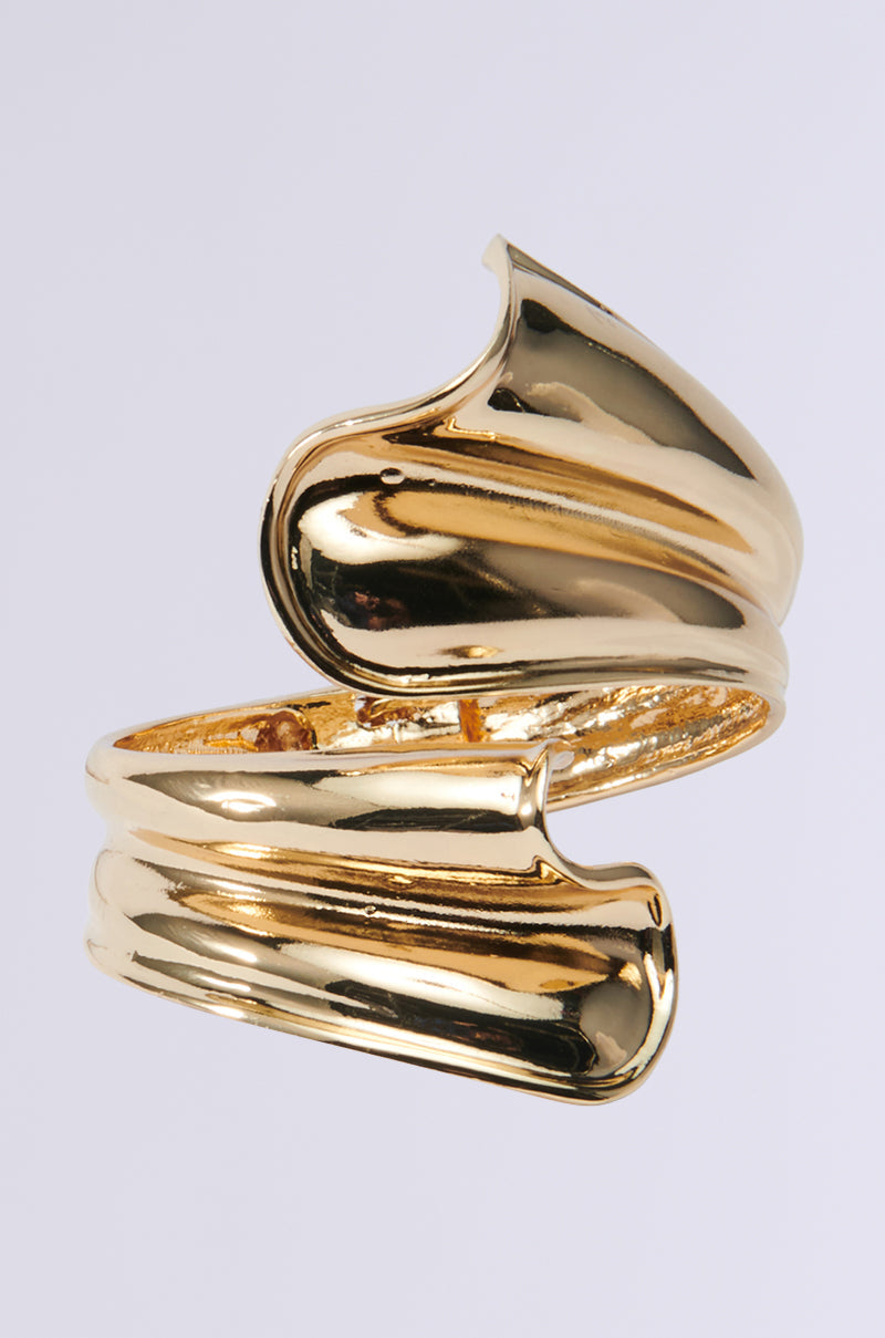 A shiny faux gold bracelet with a sculptural, twisted design against a light gray background. Its surface is smooth and reflective, showcasing intricate wraparound curves. 1