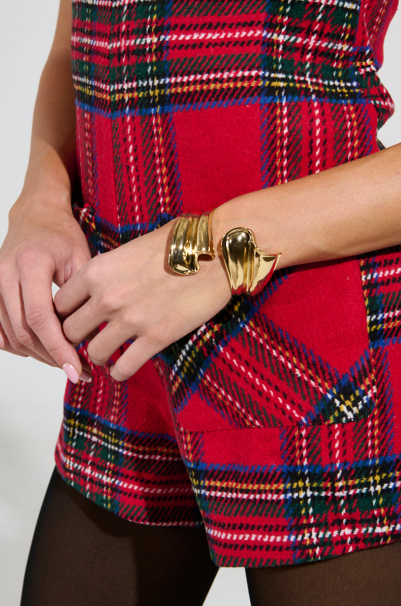 The person is wearing a red plaid outfit with short sleeves, featuring a large, shiny wraparound cuff known as the WIND IT UP BRACELET. Their ensemble highlights the checked pattern and is adorned with this faux gold bracelet, while their other hand shows off polished nails. 2