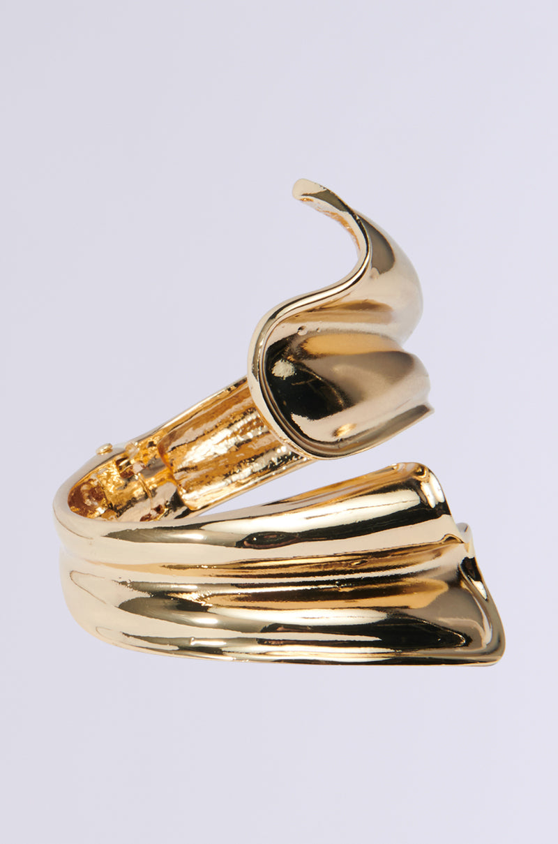 The WIND IT UP BRACELET is a shimmering, wraparound accessory with a sleek, wave-like design. This faux gold piece showcases elegantly curved, overlapping sections that exude a modern and artistic vibe against a neutral backdrop. 3