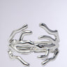 The CUFFED UP BRACELET IN SILVER features abstract, branch-like designs intertwining elegantly. This statement piece beautifully reflects light, accentuating its smooth and modern aesthetic against a plain backdrop.