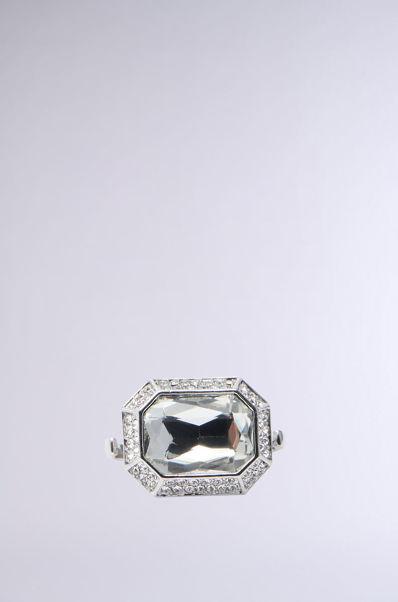 A close-up of the IN THE ESTATE RING highlights a chunky gemstone design with a large, rectangular faceted centerpiece and is elegantly encircled by a pave rhinestone trim, set against a pale gray background.