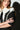 Wearing a chic black and white ensemble adorned with pearls, the individual showcases the IN THE ESTATE RING, a prominent chunky gemstone piece. Their long hair and partially shadowed appearance against a white backdrop accentuate their crossed arms and detailed attire.