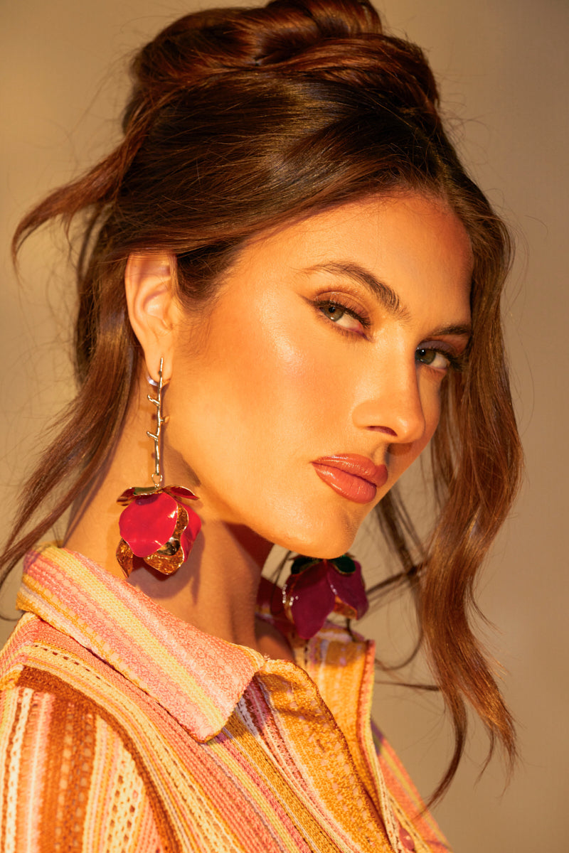 A woman with long brown hair in an updo wears IN BLOOM EARRING with a rose dangle silhouette, enhancing her profile's elegance. Her vibrant textured top in orange and pink beautifully complements the earrings' faux gold base against a neutral backdrop. 0