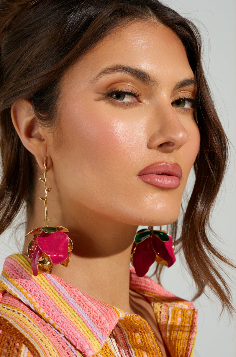 A woman with long brown hair in a loose updo wears IN BLOOM EARRING featuring large red flowers on a faux gold metallic base. She has a neutral expression and is dressed in a colorful, textured outfit against a plain, light-colored background. 2