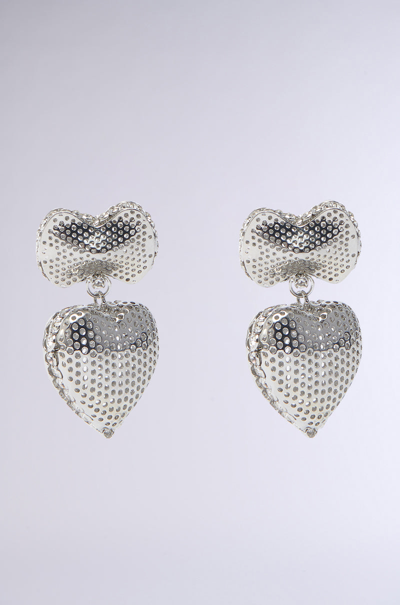 The SENDING LOVE EARRING IN SILVER features heart-shaped earrings with a textured, studded design and rhinestone trim, gracefully hanging from cloud-like forms against a light gray background.