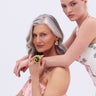 Two women pose together. The older, with long gray hair and a pink ruffled dress, sits wearing the COME TO THE GARDEN RING, while the younger blonde in a floral dress stands behind her, gently resting her arms on her shoulders.